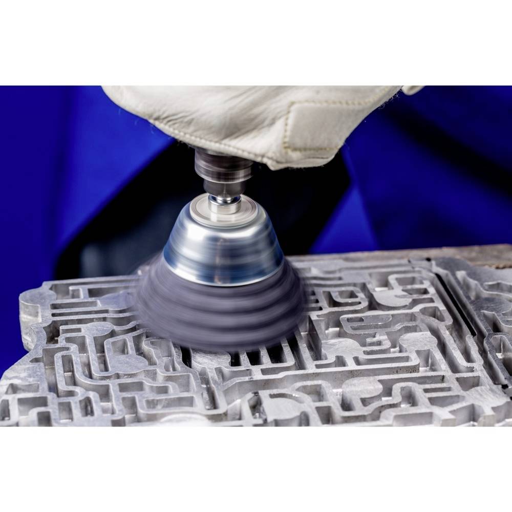 Eine Person mit Handschuh schleift ein komplexes Metallteil mit einer Maschine, wobei Funken fliegen.