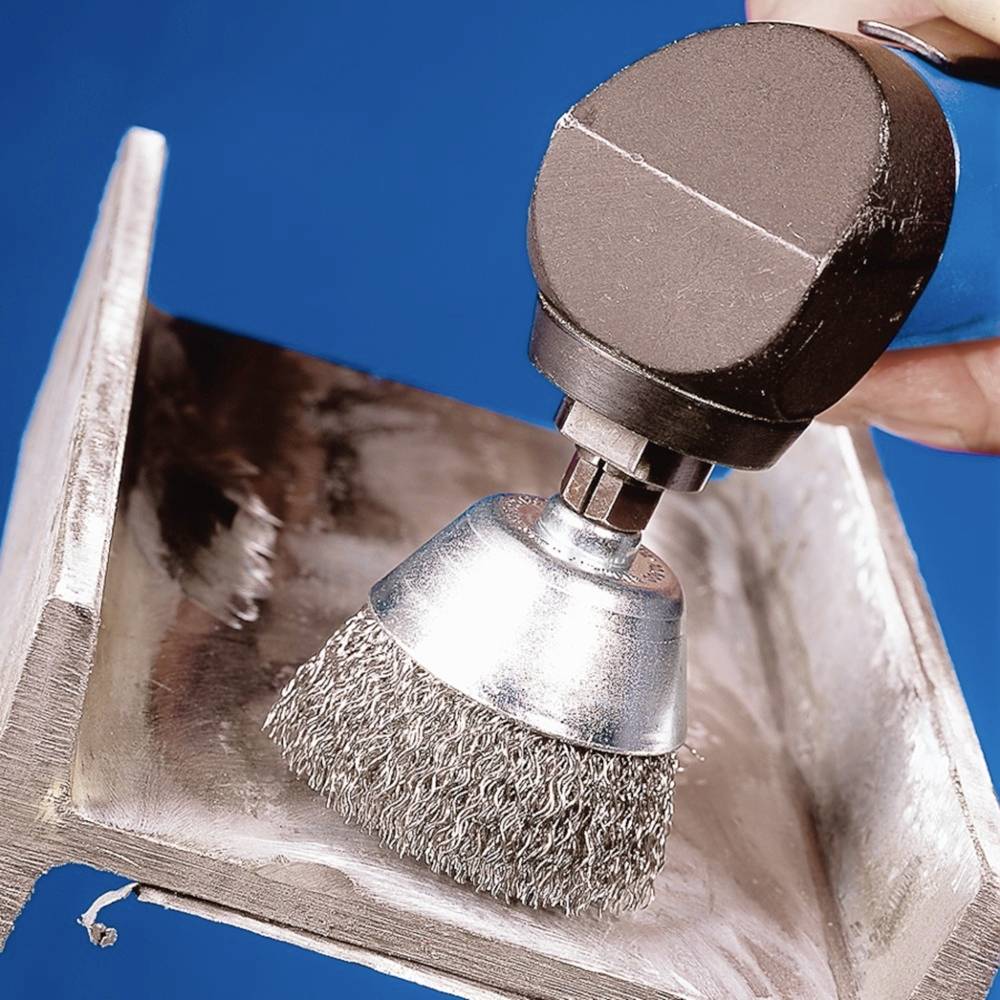 Eine Metalloberfläche wird mit einer rotierenden Drahtbürste gereinigt, um Rost zu entfernen. Hände führen das Werkzeug.