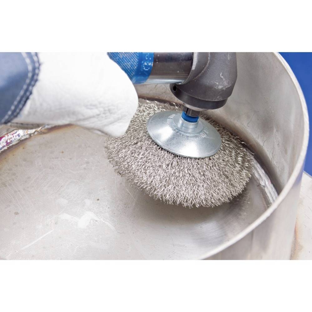 Raue Drahtbürste an elektrischer Bohrmaschine zur Reinigung einer Metalloberfläche in einer Werkstatt. Schutzhandschuh sichtbar.