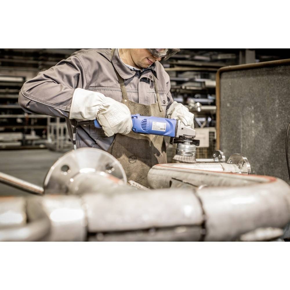 Ein Arbeiter in Schutzbekleidung schleift ein Metallrohr mit einem blauen Winkelschleifer in einer Werkstatt, umgeben von Rohrleitungen.