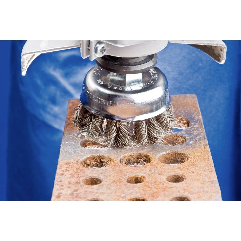 Eine Drahtbürste reinigt eine rostige Metallplatte mit Löchern. Die blauen Hintergrundtextilien heben den Fokus auf die Platte hervor.