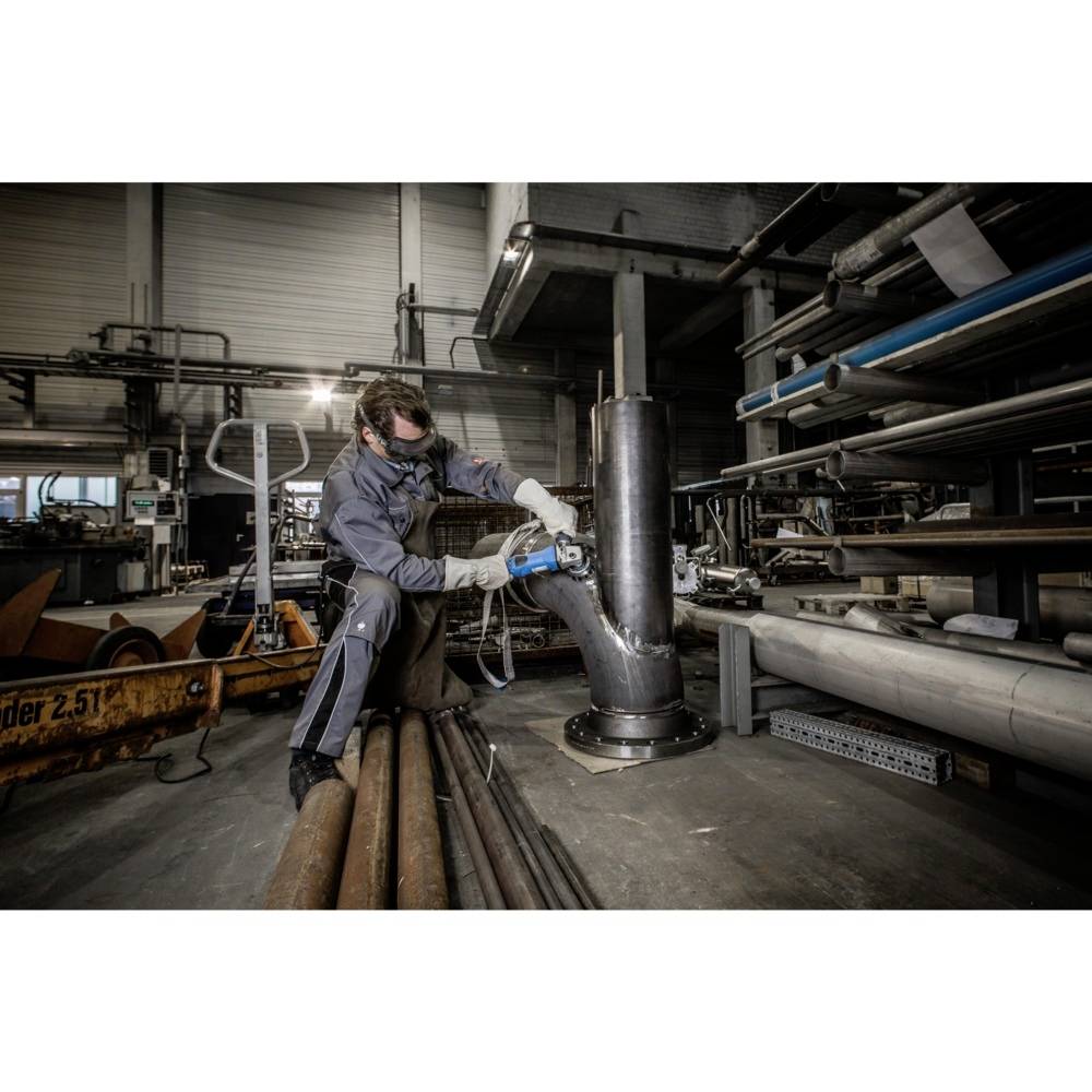 Ein Handwerker mit Schutzbrille schweißt in einer industriellen Werkstatt an einem großen Metallzylinder, umgeben von Rohren und Geräten.