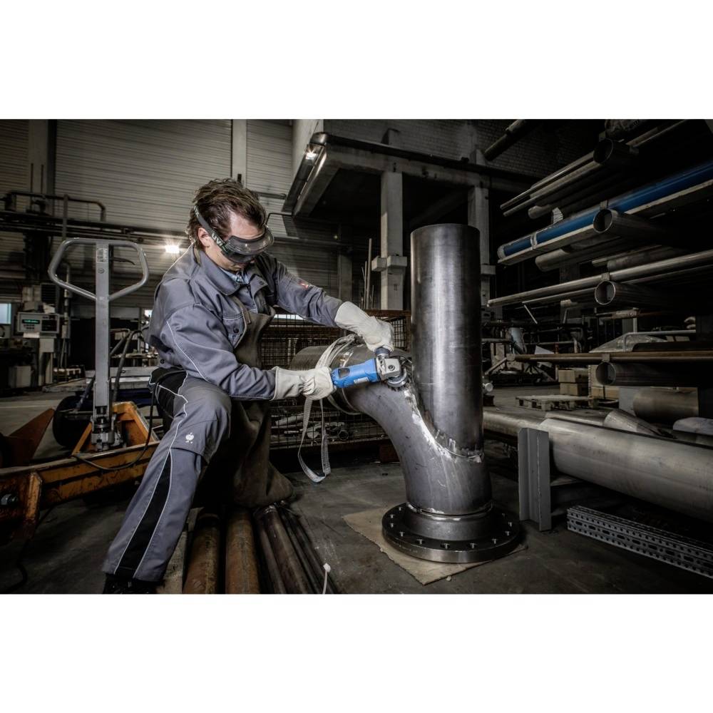 Arbeiter in Schutzkleidung schleift eine große Metallrohrverbindung in einer Werkstatt. Hintergrund zeigt verschiedene Rohre und Maschinen.