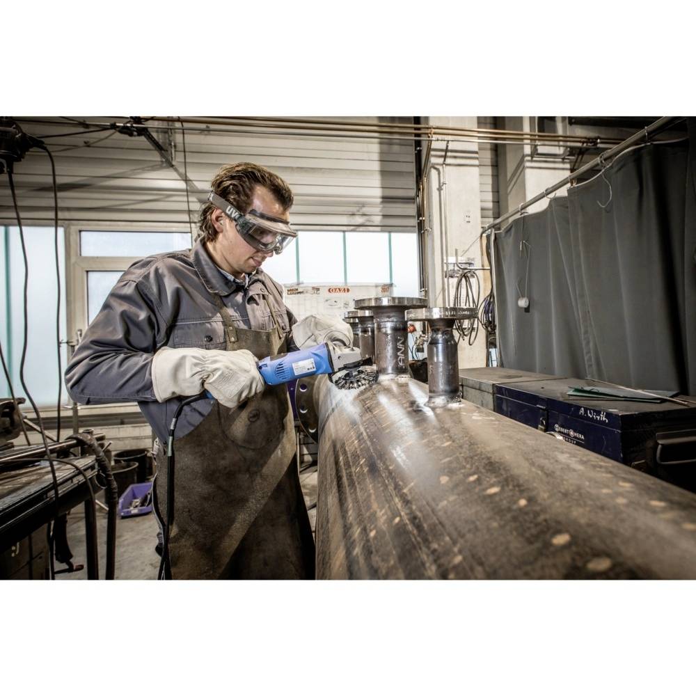 Ein Arbeiter in Schutzkleidung und Schutzbrille schleift mit einem Elektrowerkzeug an einem großen Metallzylinder in einer Werkstatt.