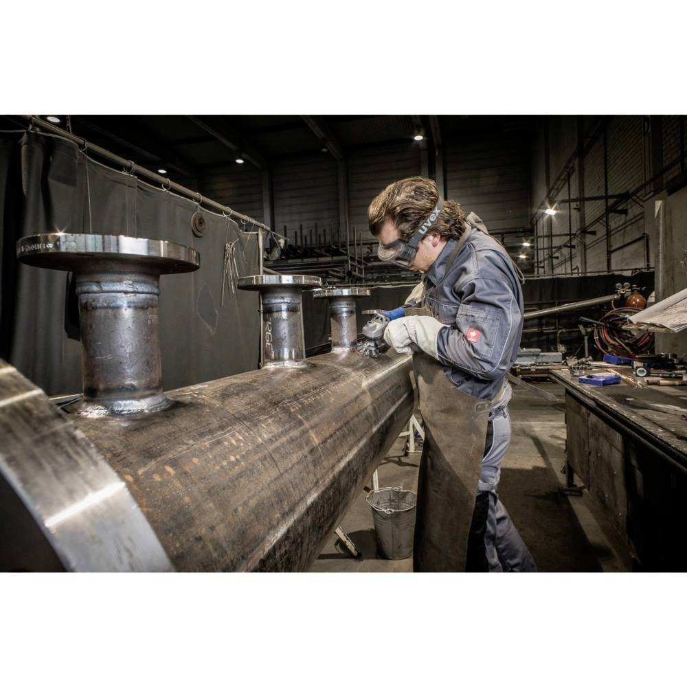 Ein Arbeiter in Schutzkleidung schweißt in einer Werkhalle ein großes Metallrohr. Umgebung wirkt industriell und gut beleuchtet.