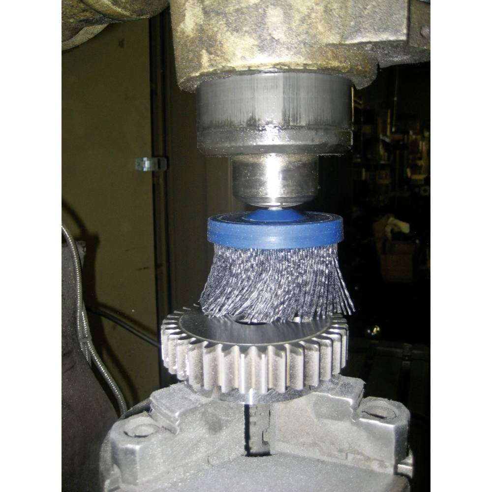 Ein Bürstenaufsatz mit blauen Borsten reinigt ein Metallzahnrad, das in einer Halterung befestigt ist. Oben ist ein mechanischer Arm sichtbar.