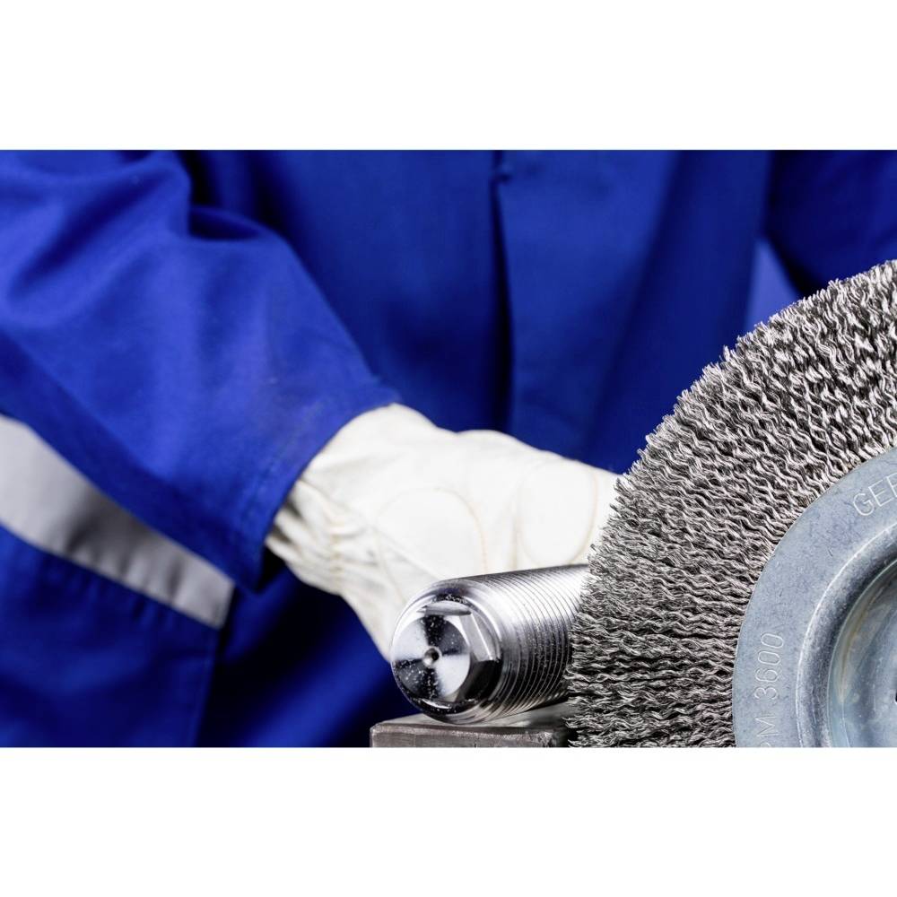 Person in blauer Arbeitskleidung hält eine rotierende Drahtbürste in einer Werkstatt. Fokus auf die Bürste und die Schutzkleidung.
