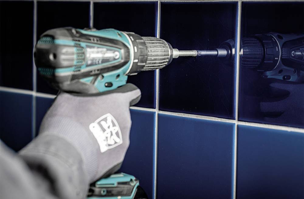 Eine behandschuhte Hand hält eine Bohrmaschine und bohrt ein Loch in eine blaue Fliese.