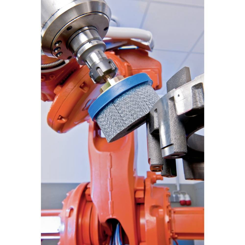 Ein Industrieroboter mit orangem Arm bearbeitet ein Werkstück mit einer rotierenden Bürste in einer Fabrikumgebung.