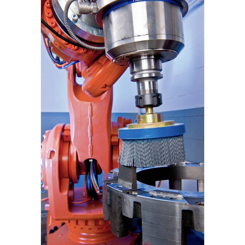 Industrieroboter, der ein Werkstück mit einer rotierenden Bürste bearbeitet. Der Roboter ist orange und befindet sich in einer Fabrikumgebung.