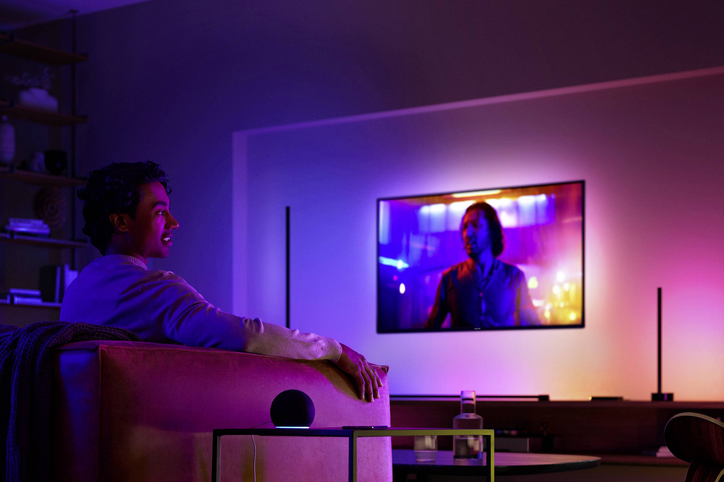 Eine Person sitzt auf einem Sofa und schaut auf einen großen Fernseher an der Wand. Der Raum ist mit bunten Lichtern beleuchtet.