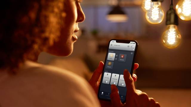 Eine Person nutzt ein Smartphone, um smarte Beleuchtung in einem Raum zu steuern. Im Hintergrund sind hängende Glühbirnen zu sehen.