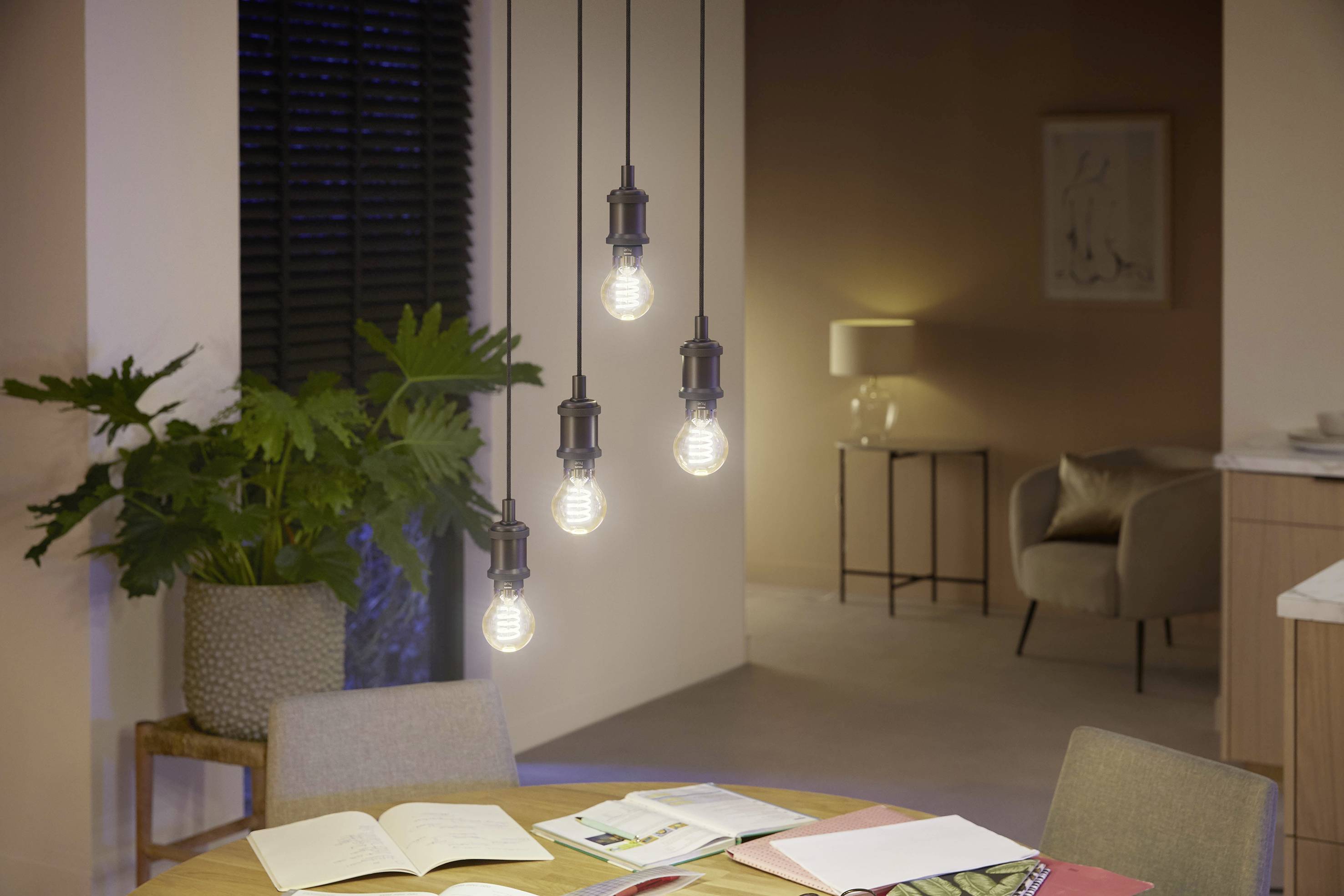 Esszimmer mit rundem Tisch und vier Hängelampen über Büchern, im Hintergrund sind ein Sessel, eine Stehlampe und eine Pflanze zu sehen.