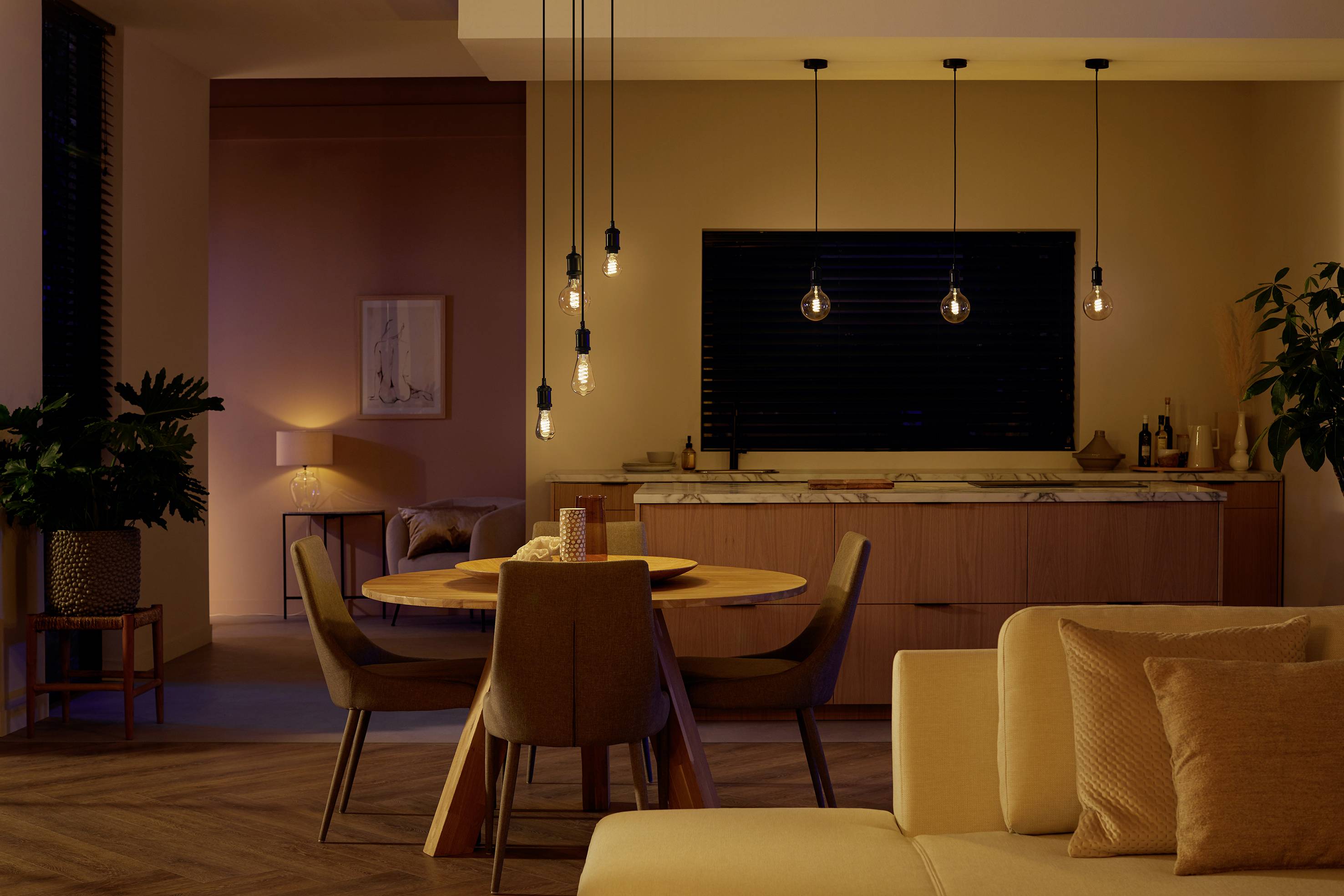 Gemütliches Wohnzimmer mit moderner Beleuchtung. Holztisch mit Stühlen, Pflanze in Ecke, Sofa im Vordergrund, gedämpfte Beleuchtung.