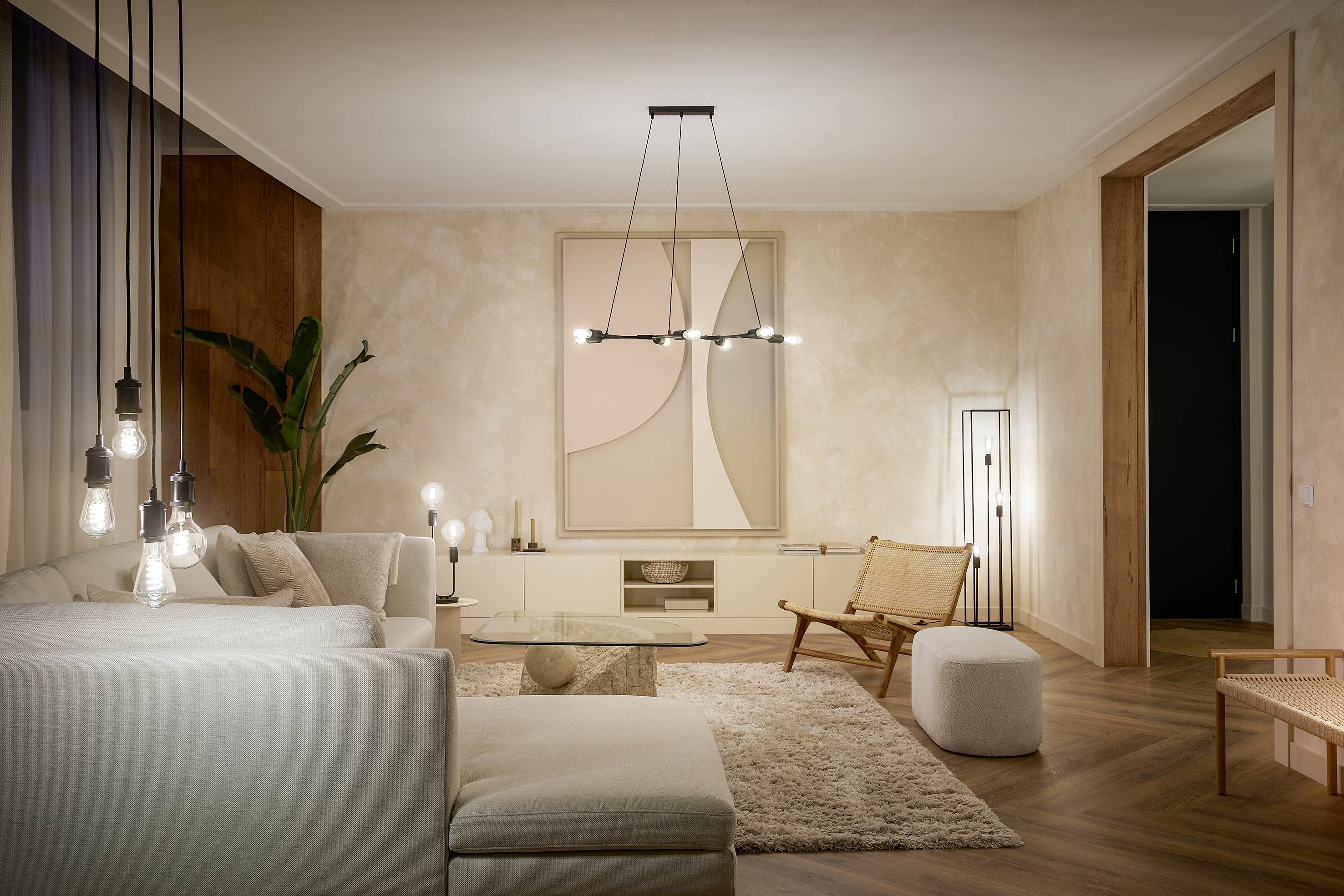 Ein modernes Wohnzimmer mit beigem Sofa, Teppich und dezentem Kunstwerk an der Wand. Pendelleuchten und Pflanzen sorgen für eine gemütliche Atmosphäre.
