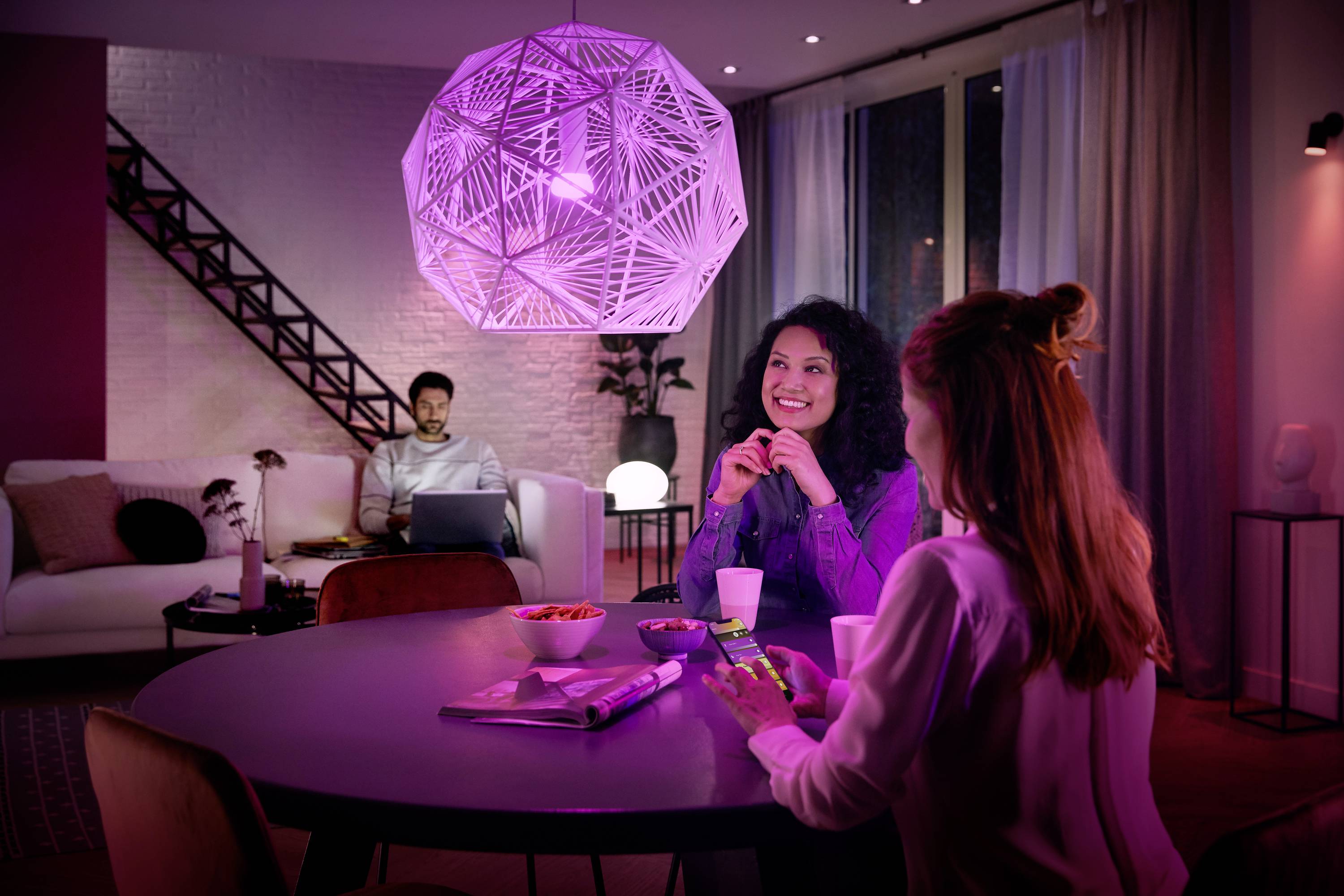 Zwei Frauen sitzen an einem Tisch unter einer großen, dekorativen Lampe und unterhalten sich. Ein Mann sitzt im Hintergrund auf einem Sofa mit einem Laptop.