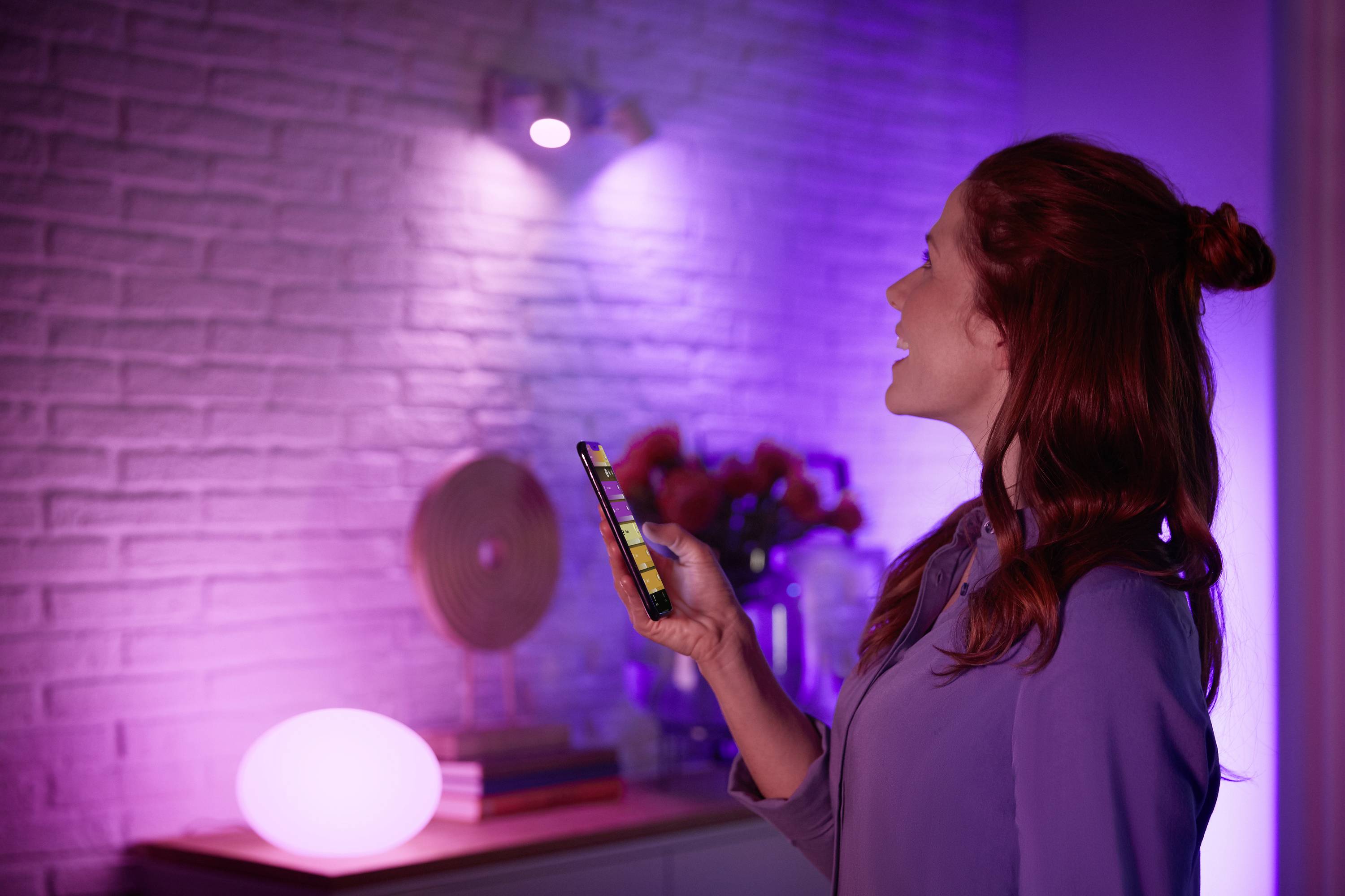 Eine Person steuert mit einem Smartphone die Beleuchtung in einem Raum, der in lila und rosa Tönen beleuchtet ist.