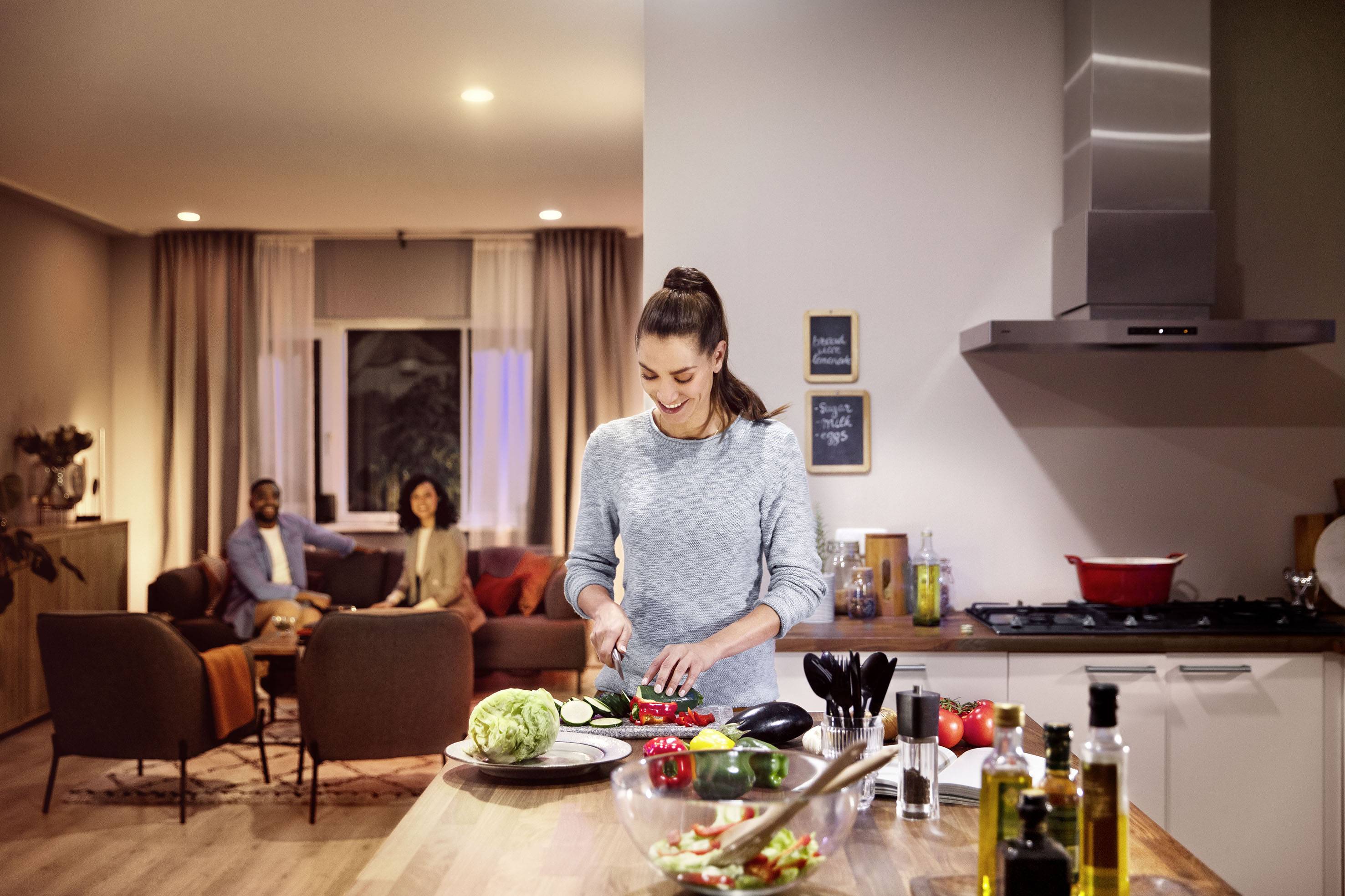 Eine Frau schneidet Gemüse in einer modernen Küche. Im Hintergrund sitzen zwei Personen auf einem Sofa und unterhalten sich.