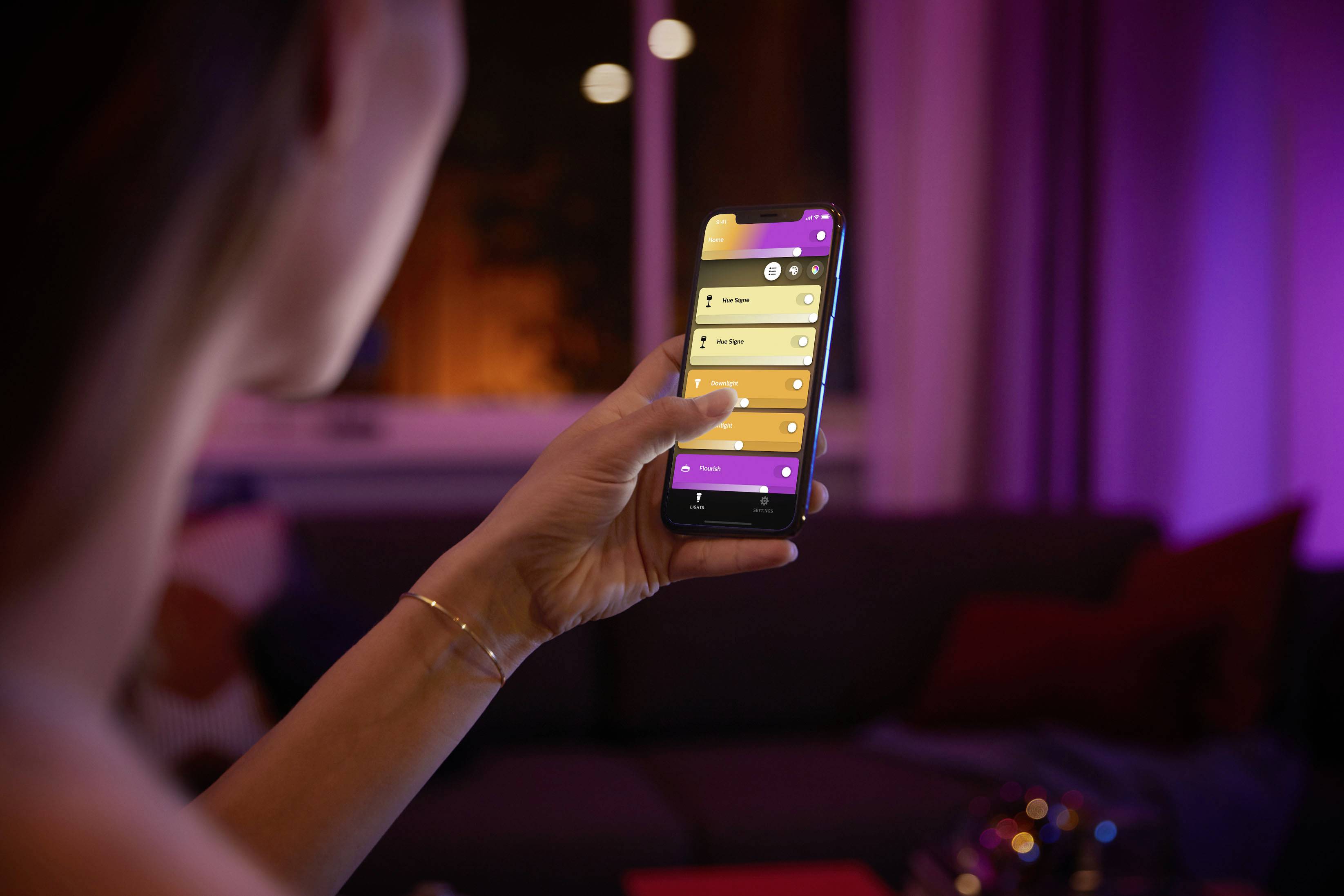 Eine Person steuert mit einem Smartphone die Beleuchtung eines Wohnzimmers, indem sie verschiedene Farb- und Helligkeitsoptionen auswählt.