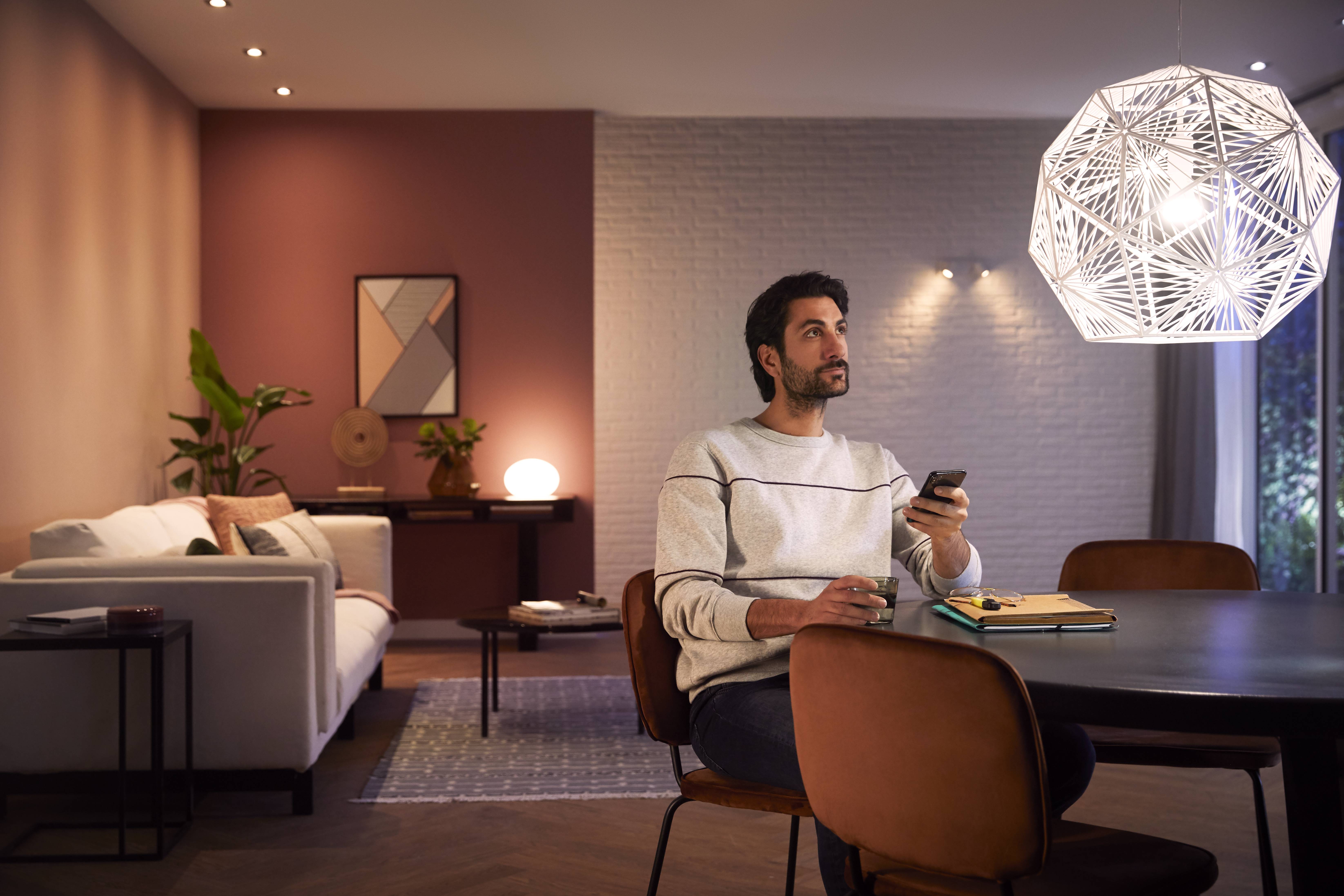 Ein Mann sitzt am Esstisch in einem modern beleuchteten Wohnzimmer, schaut nachdenklich und hält eine Fernbedienung in der Hand.