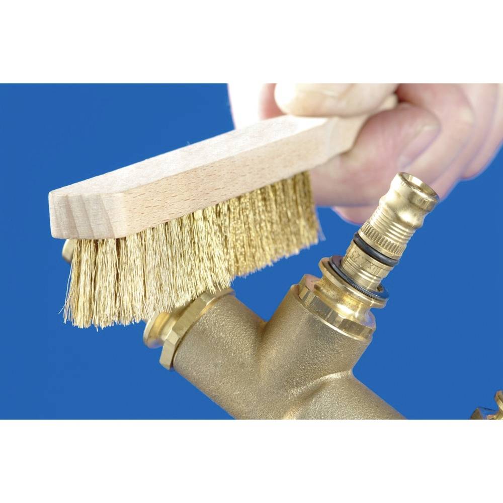 Eine Hand hält eine Drahtbürste und reinigt ein Messingrohr vor blauem Hintergrund.