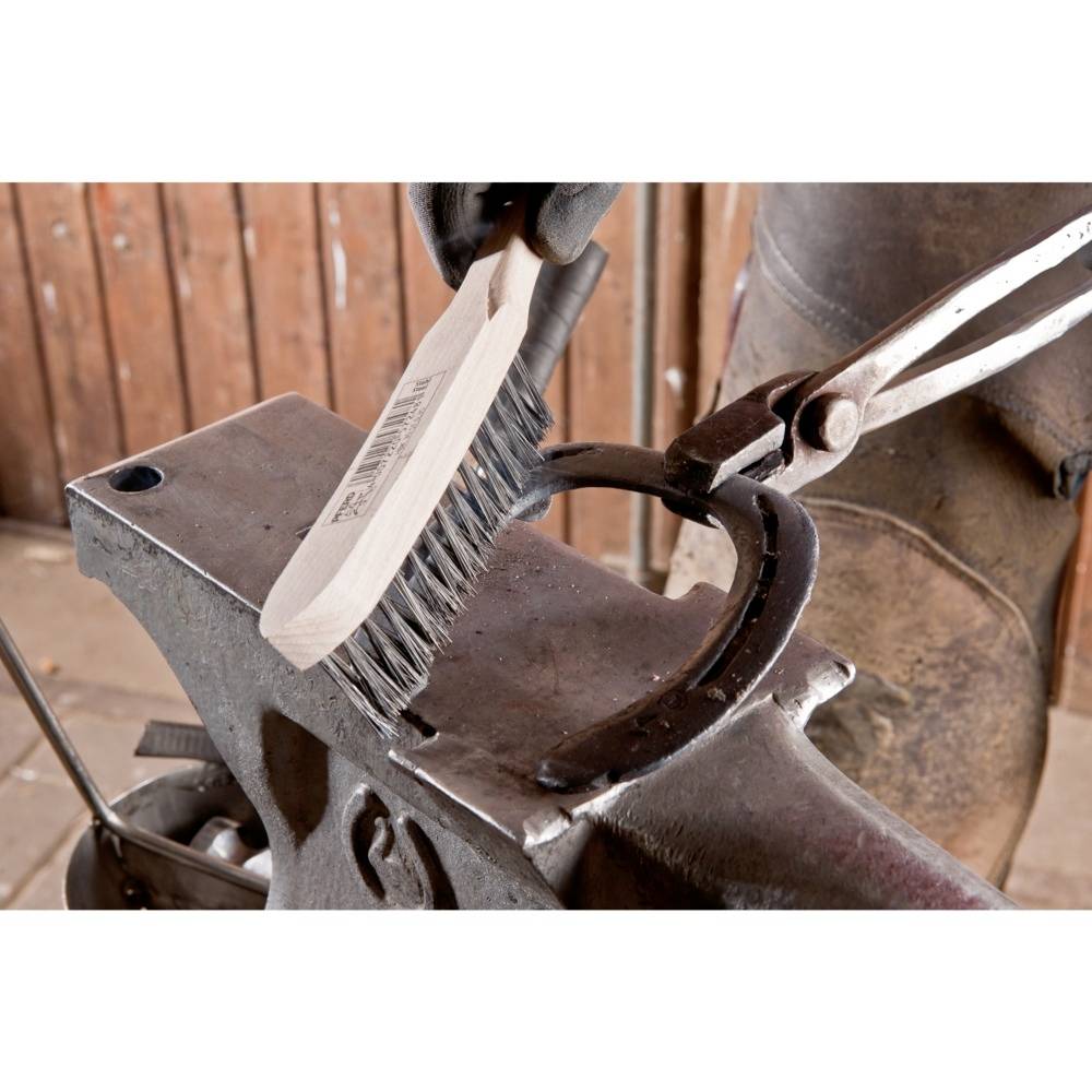 Eine Hand reinigt einen Amboss mit einer Drahtbürste. Im Hintergrund ist eine Holzwand sichtbar.
