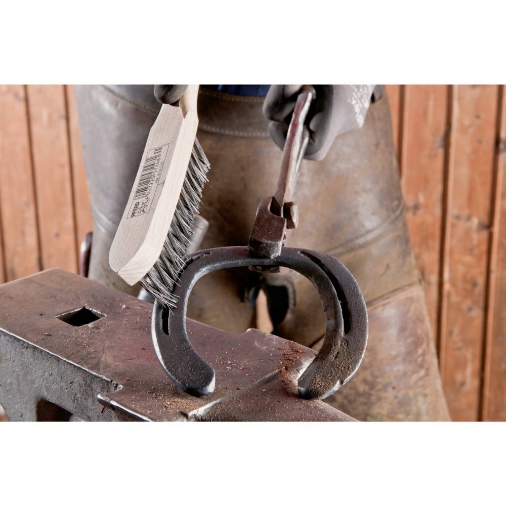 Eine Person, die in Arbeitskleidung ein Hufeisen mit einer Drahtbürste reinigt. Das Hufeisen liegt auf einem Amboss.