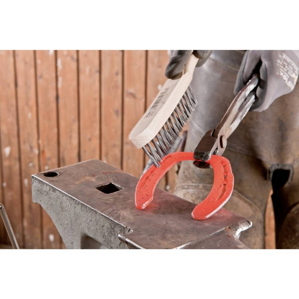 Eine Person hält ein Hufeisen mit einer Zange und reinigt es mit einer Drahtbürste auf einem Amboss. Im Hintergrund Holzverkleidung.