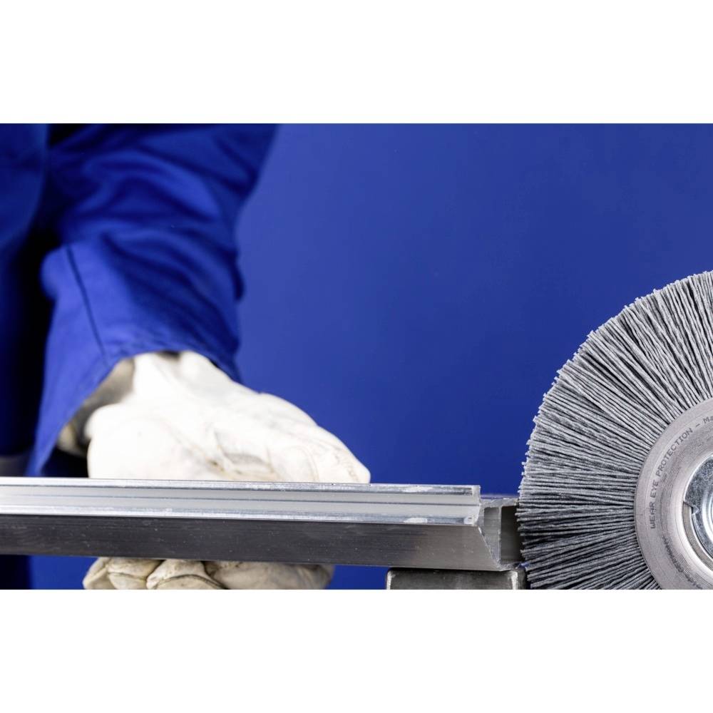 Eine Person in blauer Arbeitskleidung hält ein Metallwerkstück neben einer rotierenden Drahtbürste, die für Abreibearbeiten verwendet wird.