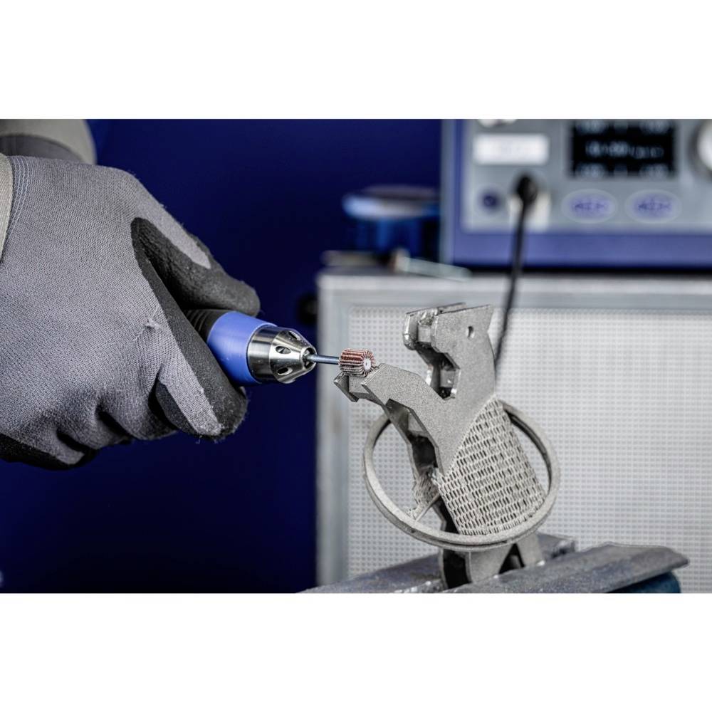 Eine Person bearbeitet ein 3D-gedrucktes Objekt mit einem rotierenden Werkzeug, das graue Material entfernt. Im Hintergrund steht ein Gerät.