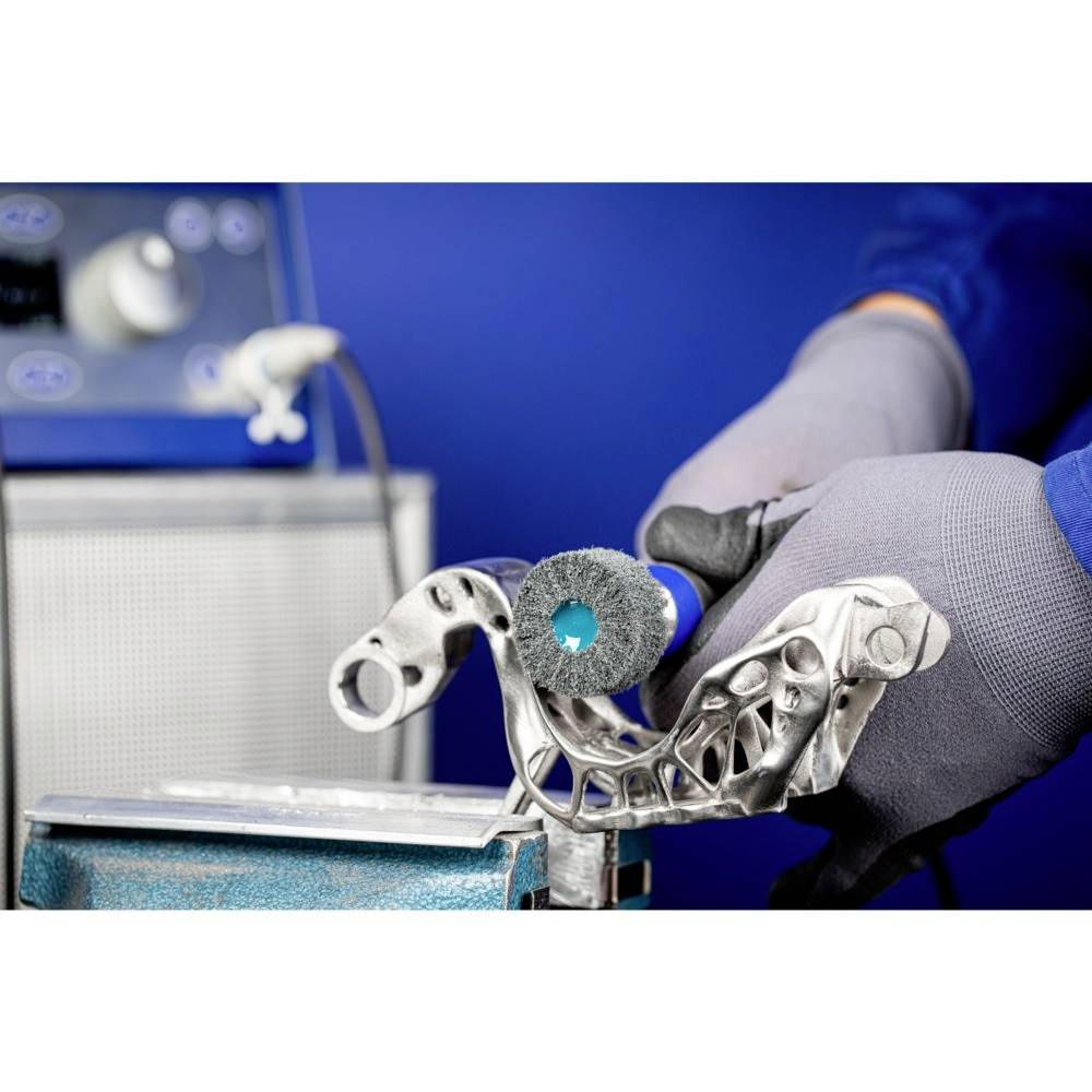 Eine gloved Hand verwendet ein rotierendes Schleifwerkzeug, um Details an einem komplexen Metallwerkstück zu bearbeiten. Im Hintergrund ist ein unscharfes Gerät sichtbar.