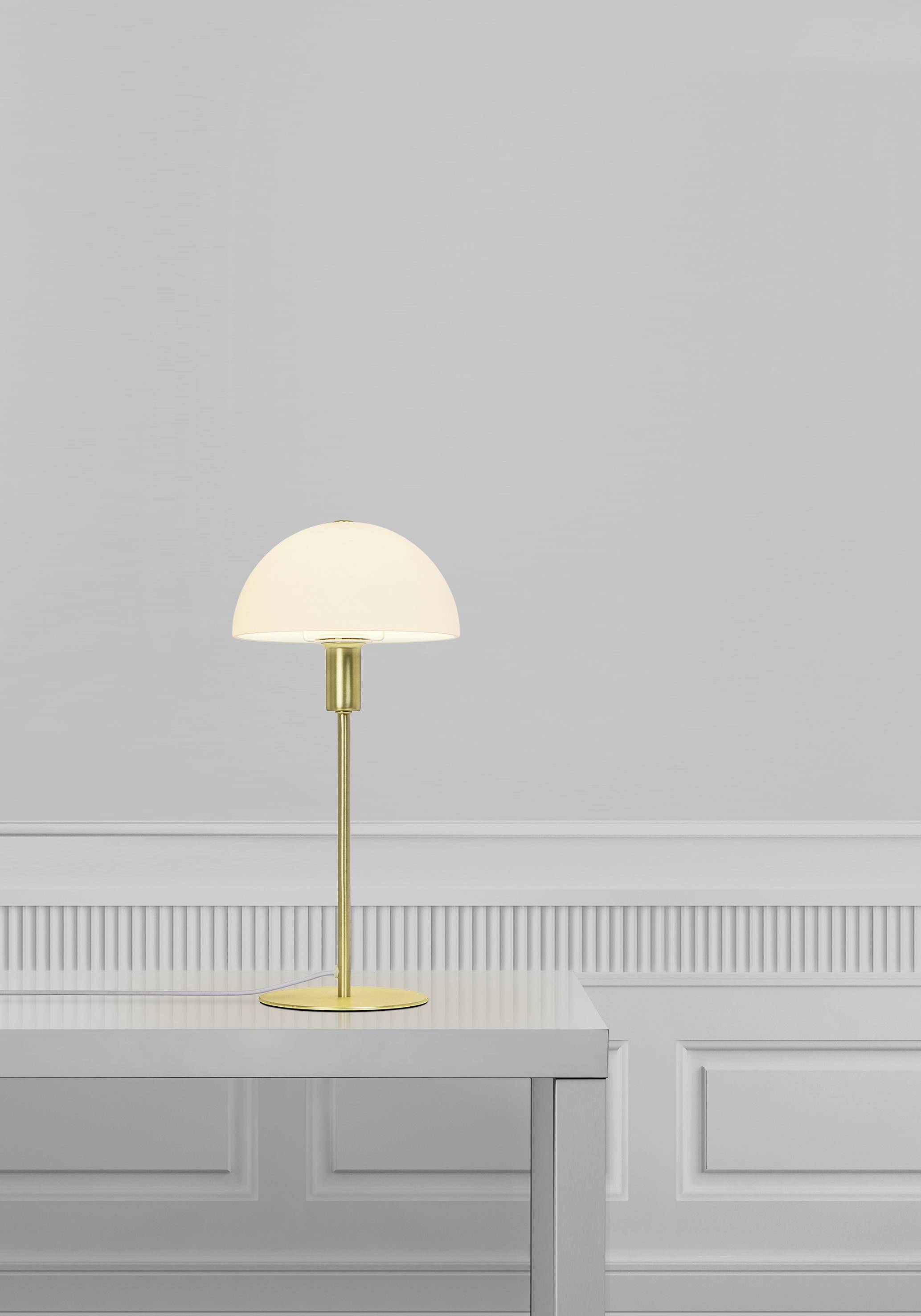Eine minimalistische Tischlampe mit goldenem Ständer und weißem Schirm steht auf einem schlichten weißen Tisch vor einer grauen Wand.