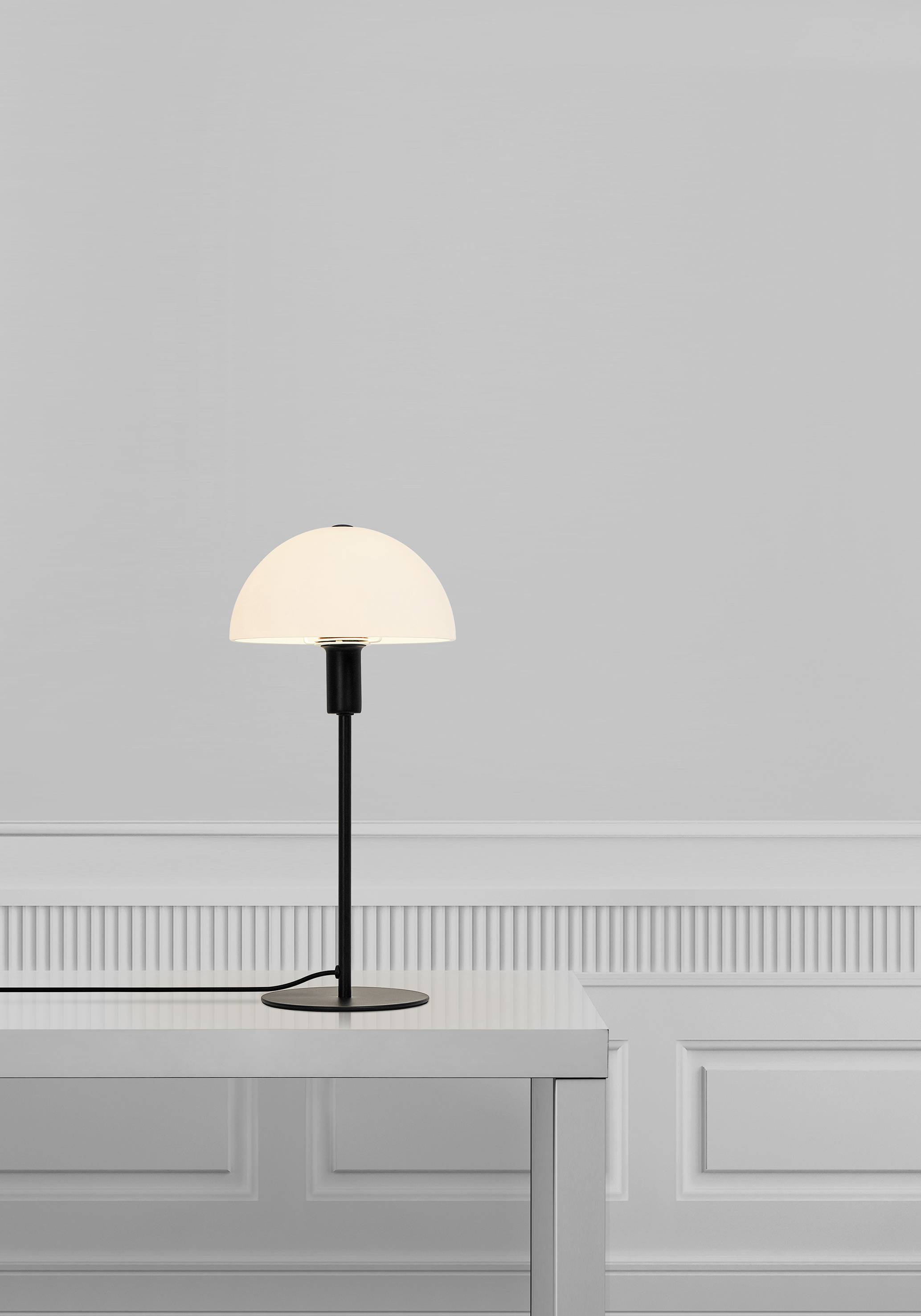 Eine minimalistische Tischlampe mit rundem, weißem Schirm steht auf einem weißen Tisch vor einer hellgrauen, schlichten Wand.