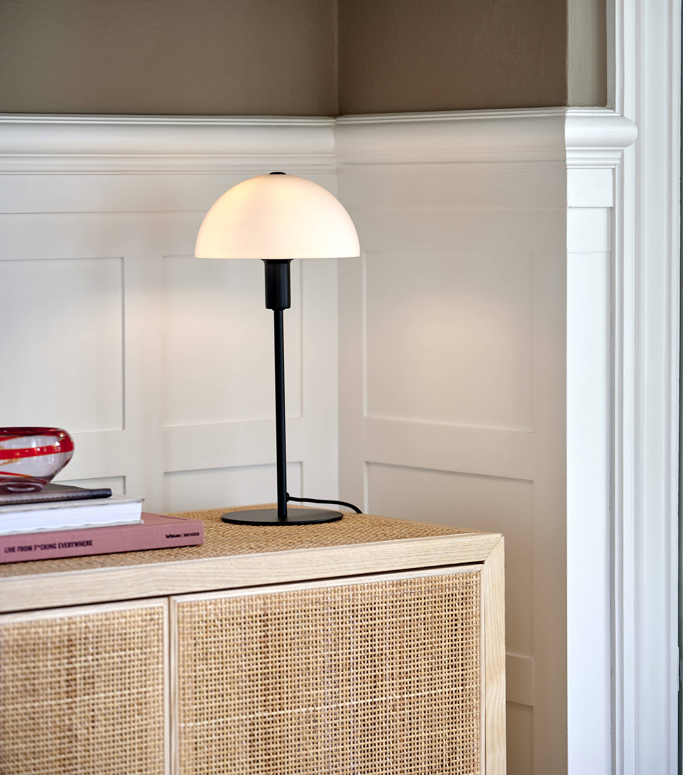 Tischlampe mit halbrundem Schirm steht auf einem Sideboard. Daneben liegen Bücher und eine rote Glasdekoration.