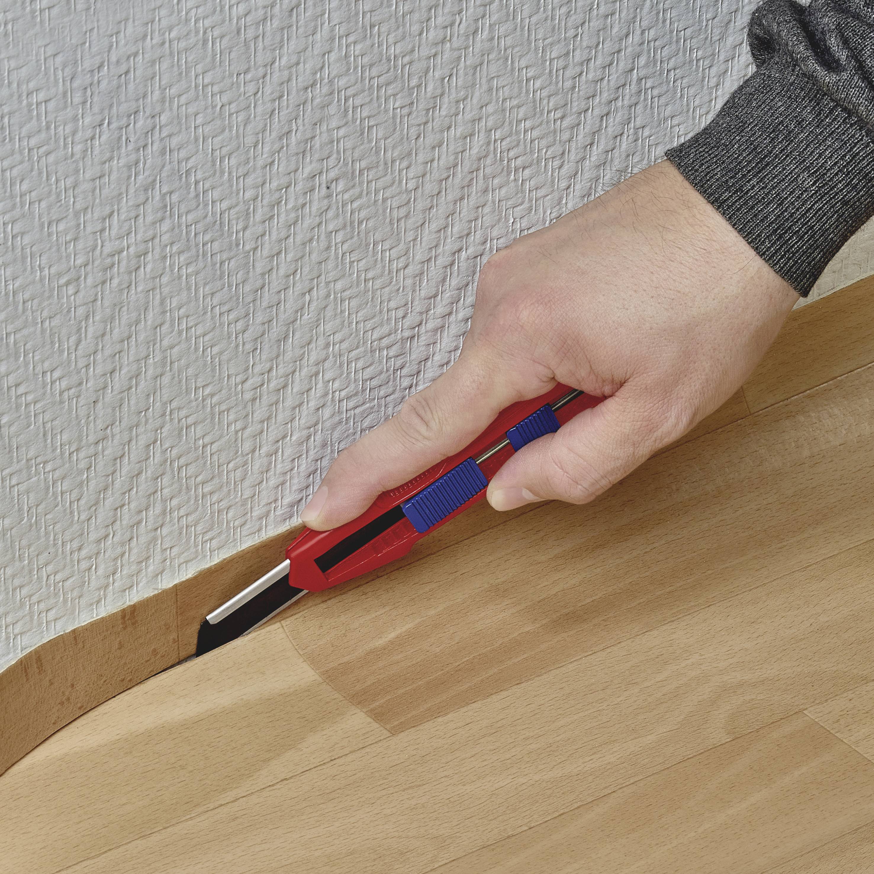 Eine Hand schneidet mit einem Cuttermesser ein Stück Teppich entlang eines Bodenbelags ab.