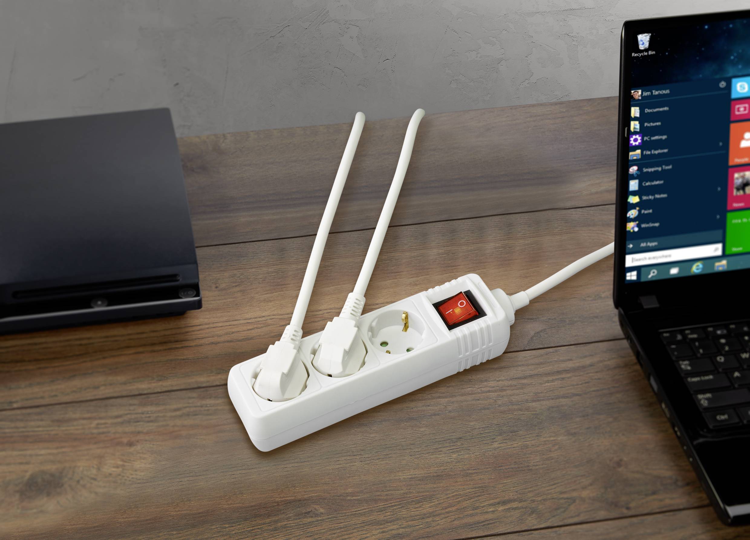 'Mehrfachsteckdose mit zwei eingesteckten Kabeln neben einem Laptop auf einem Holztisch. Der Schalter der Steckdose ist auf 'Ein'.'