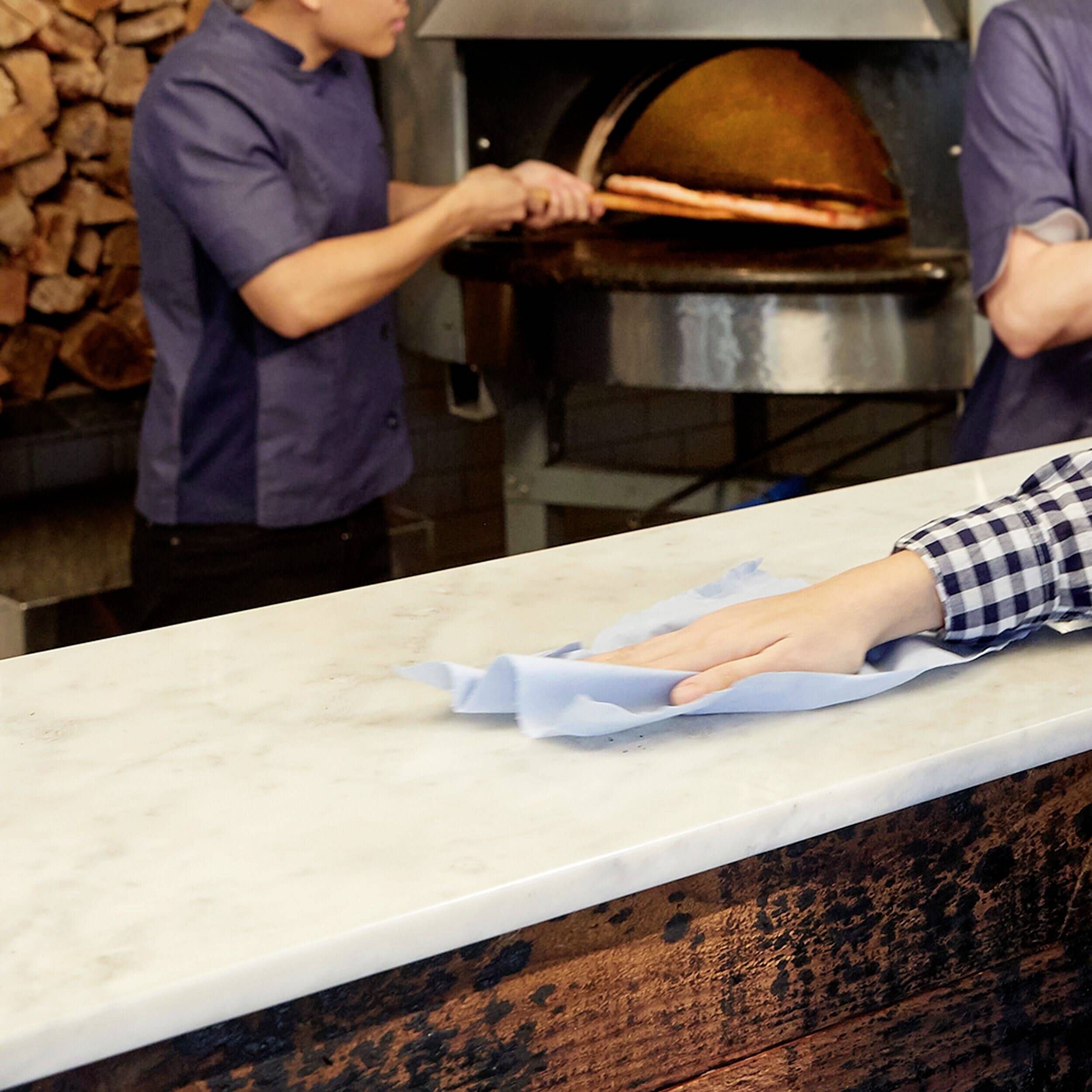 Zwei Personen in einer Bäckerei. Eine Person backt Brot im Ofen, während die andere den Tresen mit einem Tuch reinigt.