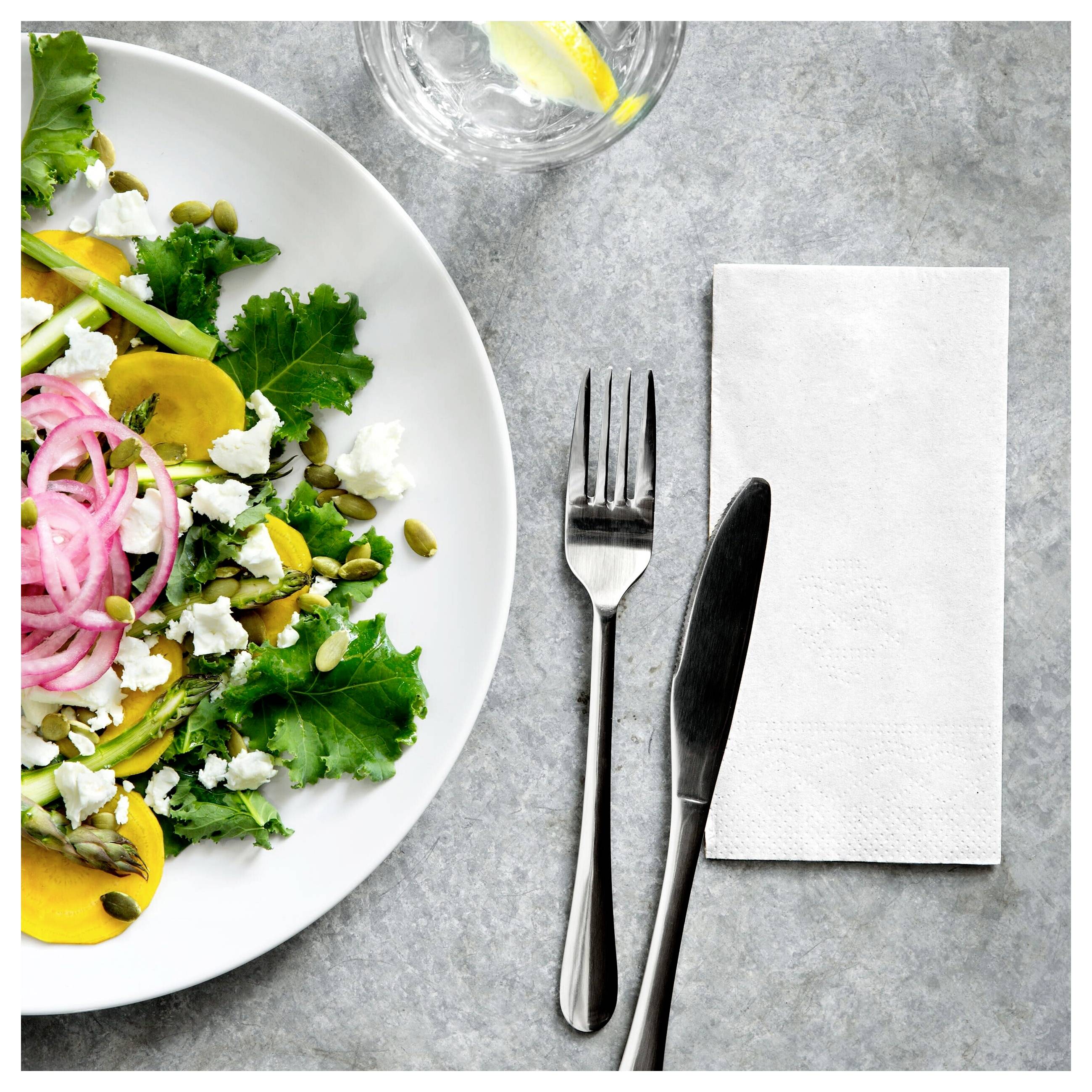 Draufsicht auf einen angerichteten Salat mit Blattgemüse, eingelegten Zwiebeln und Saaten neben einer Gabel, einem Messer, einer Serviette und einem Trinkglas auf einem grauen Tisch.
