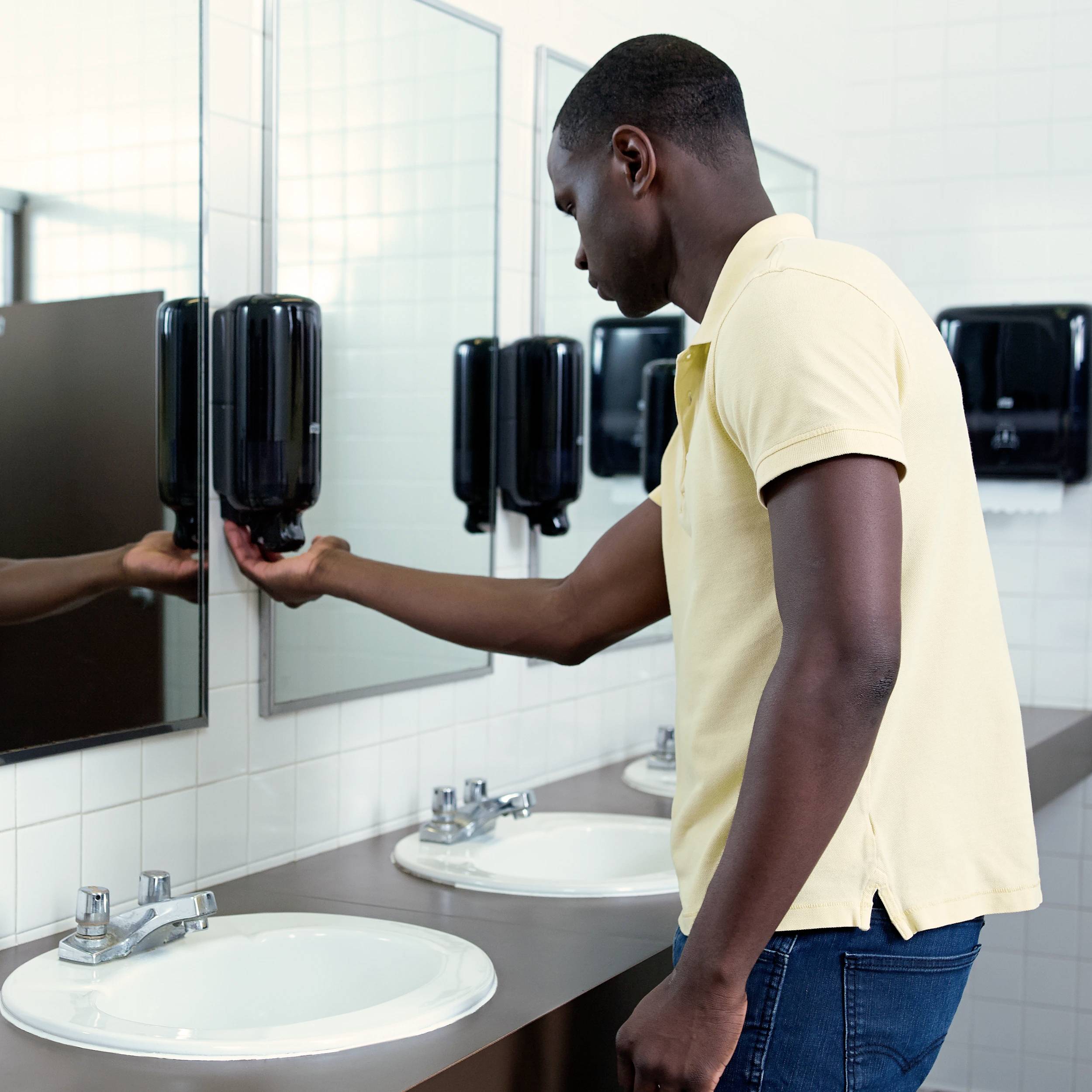 Ein Mann steht in einem Badezimmer vor einem Spiegel und benutzt einen Seifenspender. An der Wand sind weitere Waschbecken zu sehen.