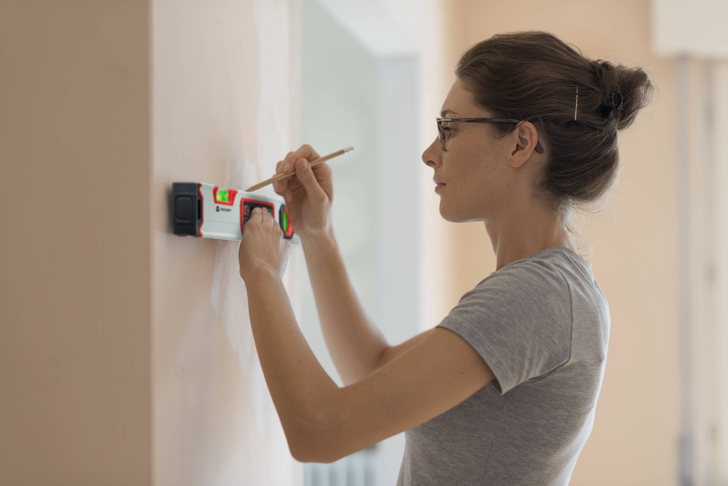 Eine Frau benutzt ein elektronisches Messgerät an einer Wand, während sie mit einem Stift eine Markierung setzt. Sie trägt ein graues T-Shirt.