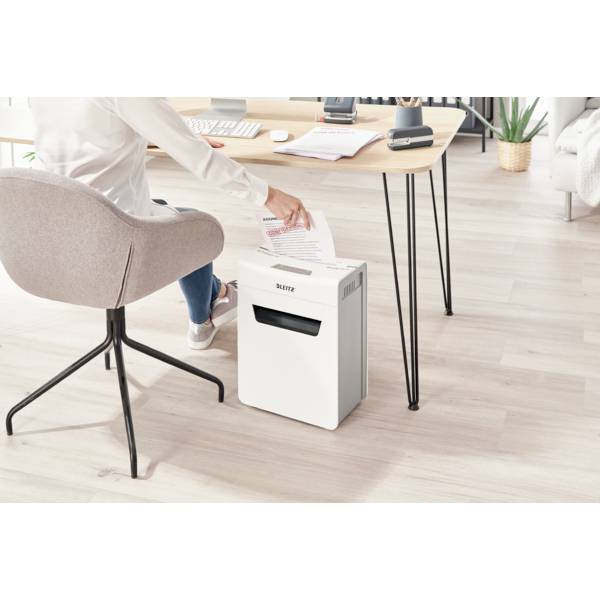 Person sitzt am Schreibtisch und steckt ein Papier in einen Aktenvernichter. Büro-atmosphäre mit Holzboden und moderner Einrichtung.