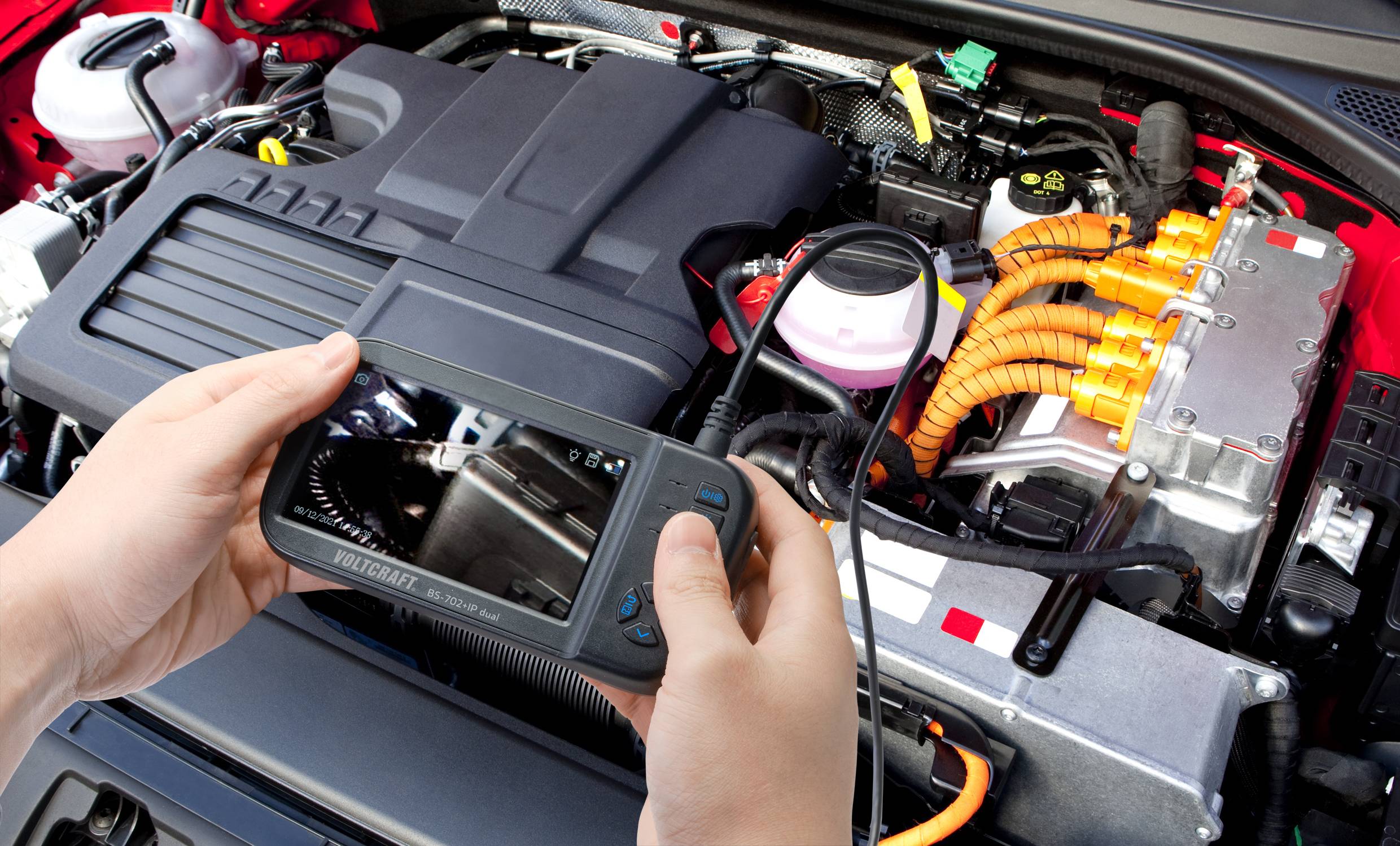 Person hält Diagnosegerät über Motorraum eines Autos. Oranger Kabelstrang und andere Bauteile sind sichtbar. Szene zeigt Wartungssituation.