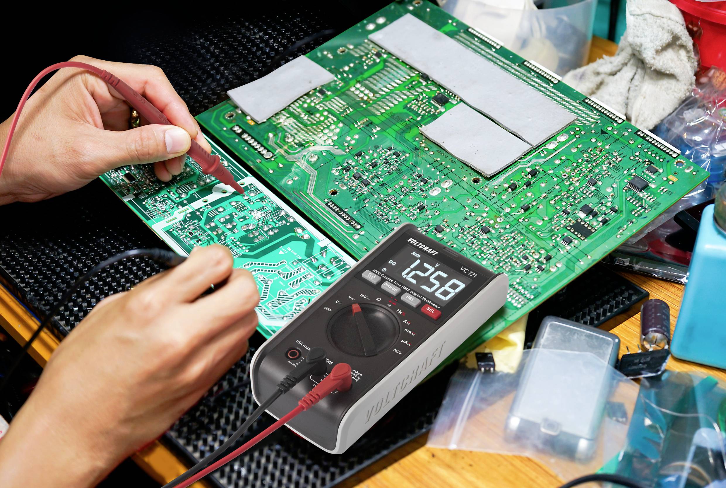 Eine Person testet eine grüne Leiterplatte mit einem Multimeter. Das Display zeigt '12.58'. Umgeben von Elektronik und Werkzeugen.