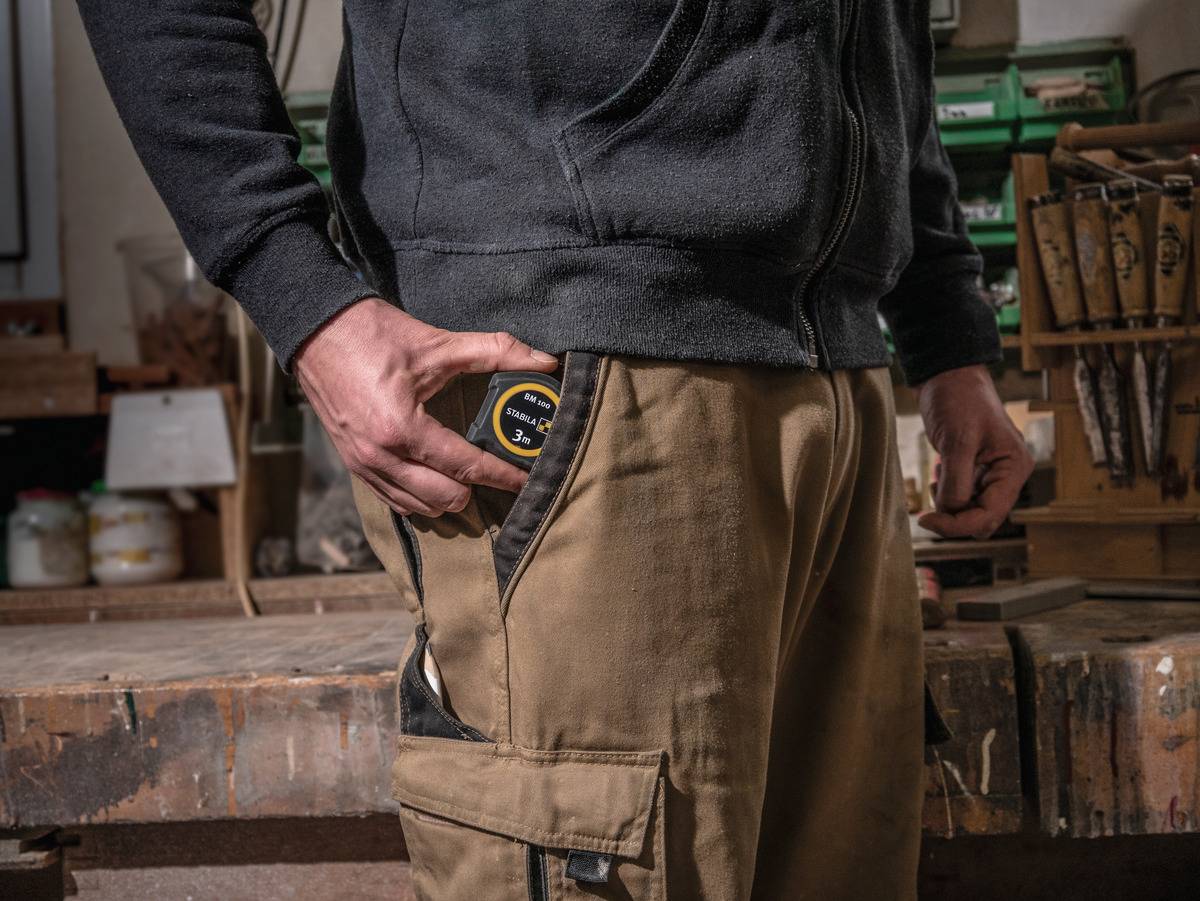 Eine Person steckt ein Maßband in die Tasche einer Werkhose in einer Werkstatt. Tisch mit Werkzeugen im Hintergrund.