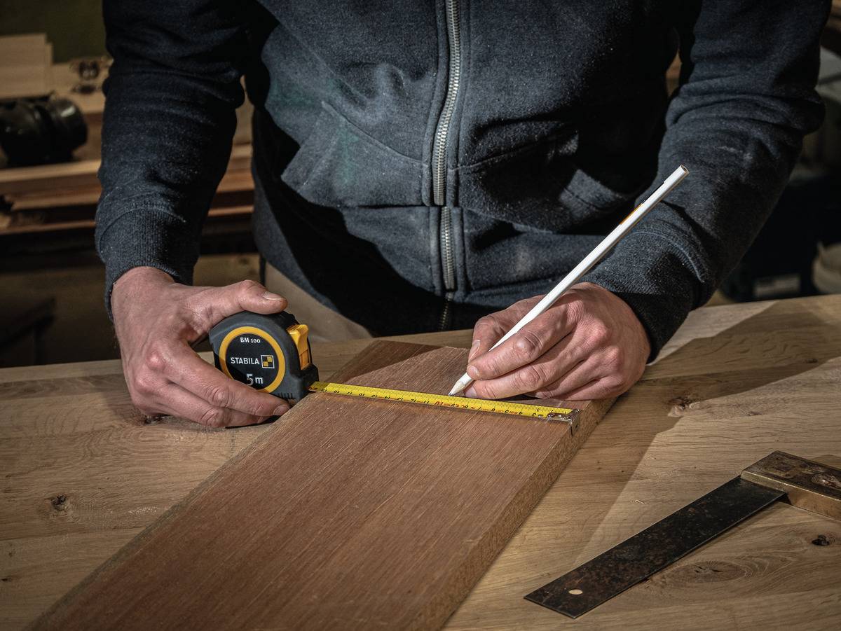 Eine Person misst ein Holzbrett mit einem Maßband und markiert es mit einem Bleistift. Ein Winkelmesser liegt daneben.