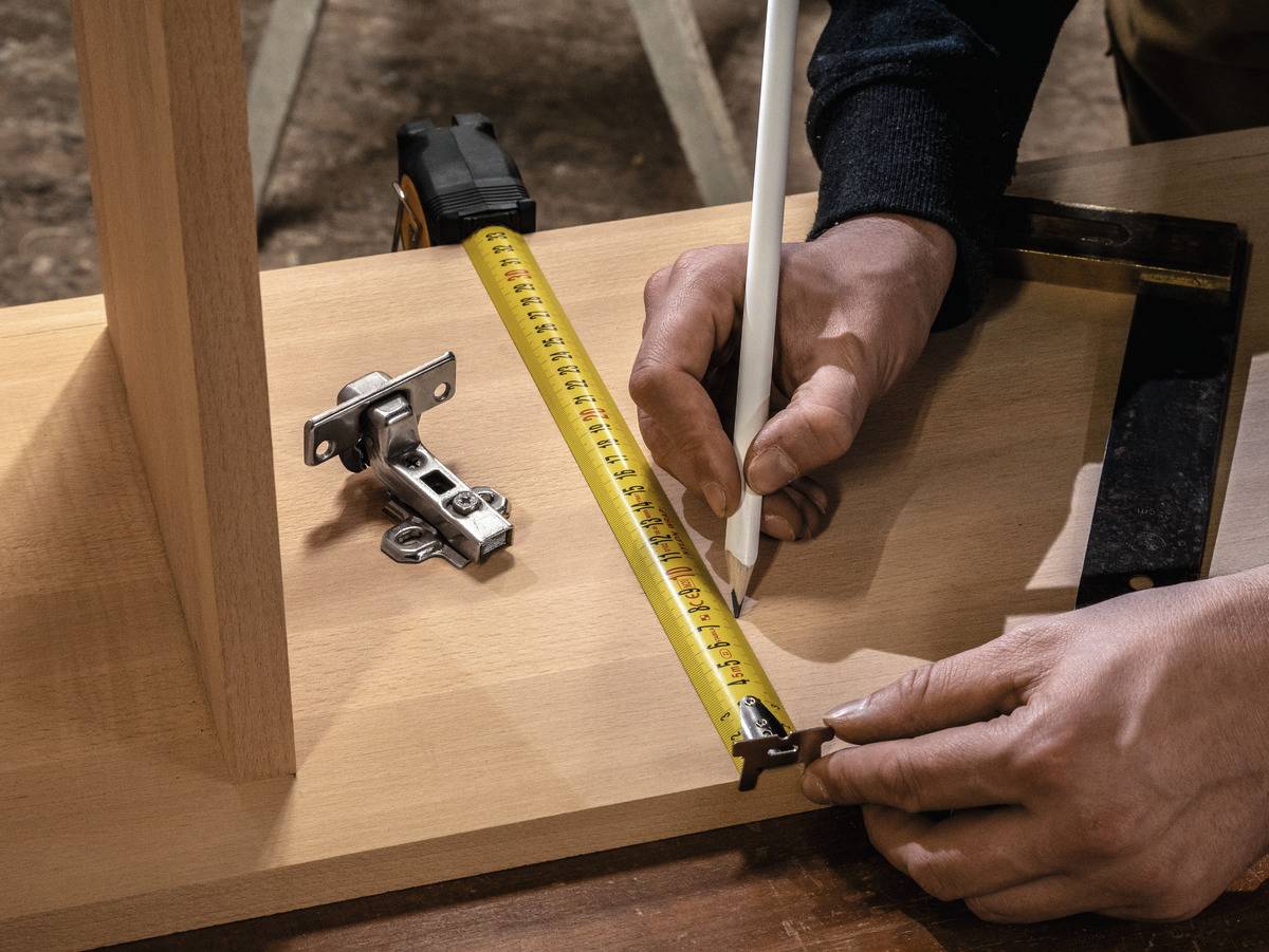 Eine Person misst mit einem Meterstab die Länge eines Holzbretts und markiert es mit einem Bleistift.