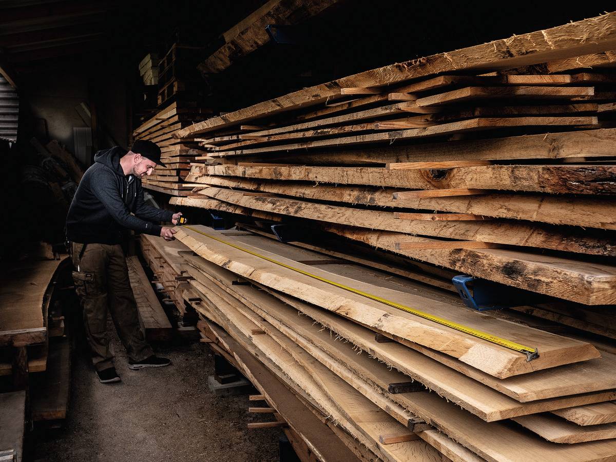 Ein Mann misst mit einem Maßband ein langes Holzbrett in einer Holzwerkstatt, umgeben von gestapelten Holzbrettern.