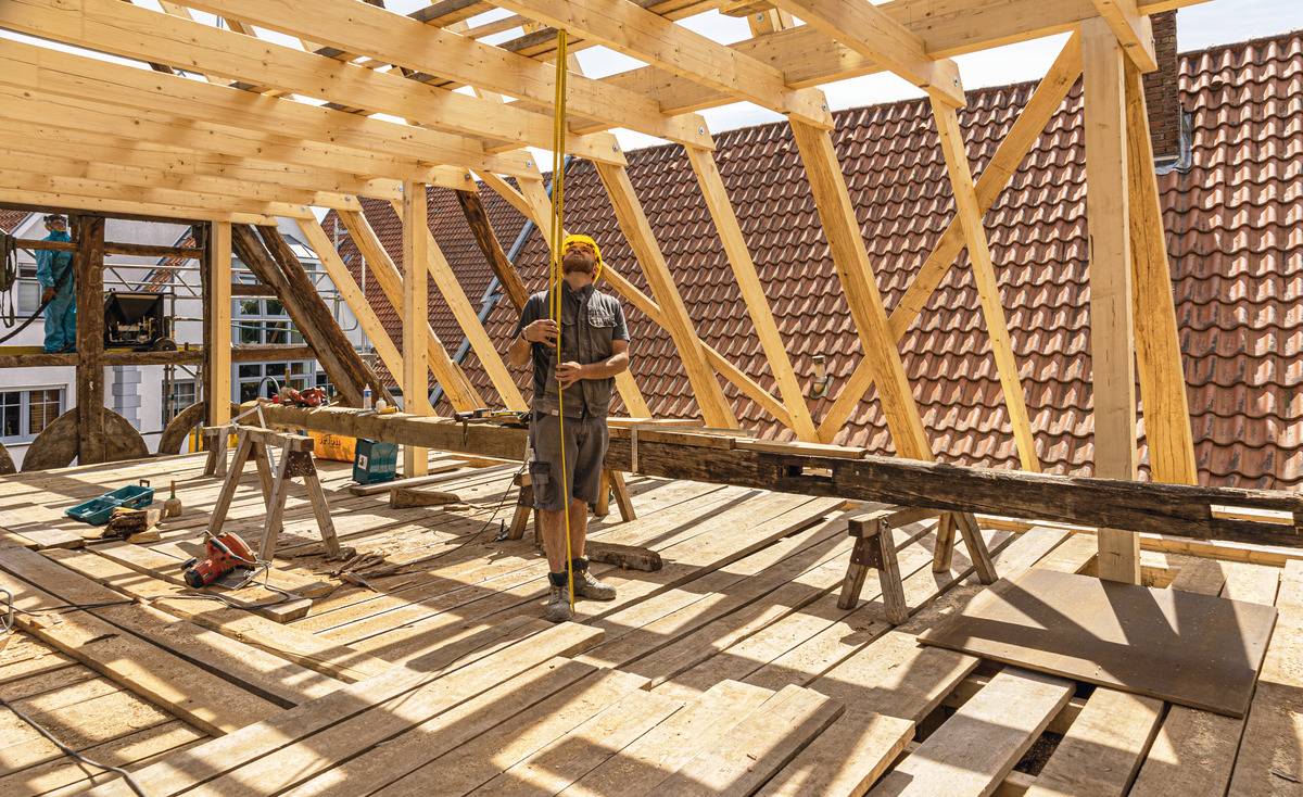 Eine Person arbeitet auf einer Baustelle beim Bau eines Holzhausdaches, umgeben von Holzbalken und Geräten. Dachziegel im Hintergrund.