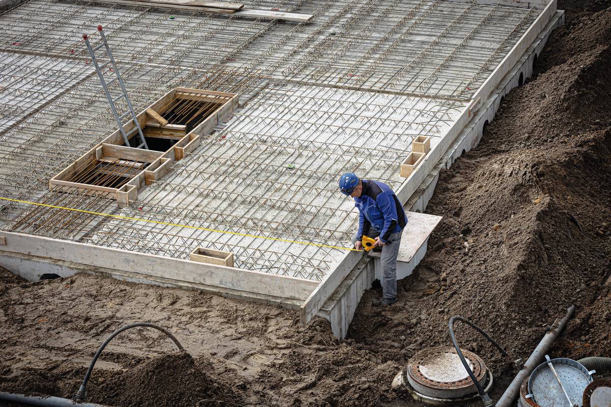 Ein Bauarbeiter misst die Betonsohlen eines Gebäudefundaments mit einem Maßband. Eisenstrukturen und Werkzeuge sind sichtbar.