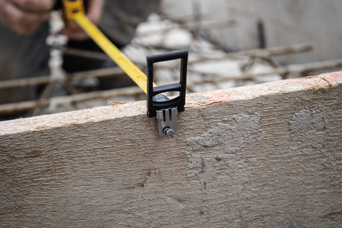 Eine Person misst ein Holzbrett mit einem Maßband auf einer Baustelle. Im Hintergrund sind Betoneisen erkennbar.